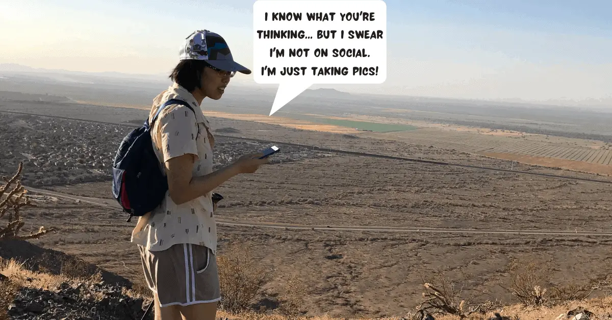 Meggie is holding up her smartphone and looking at the screen. She says "I know what you're thinking... but I swear i'm not on social. i'm just taking pics!" She is on top of a desert mountain. Below her is a dry valley of desert, a lonely highway, and a small suburban neighborhood. Phoenix, Arizona's South Mountain park, United States.