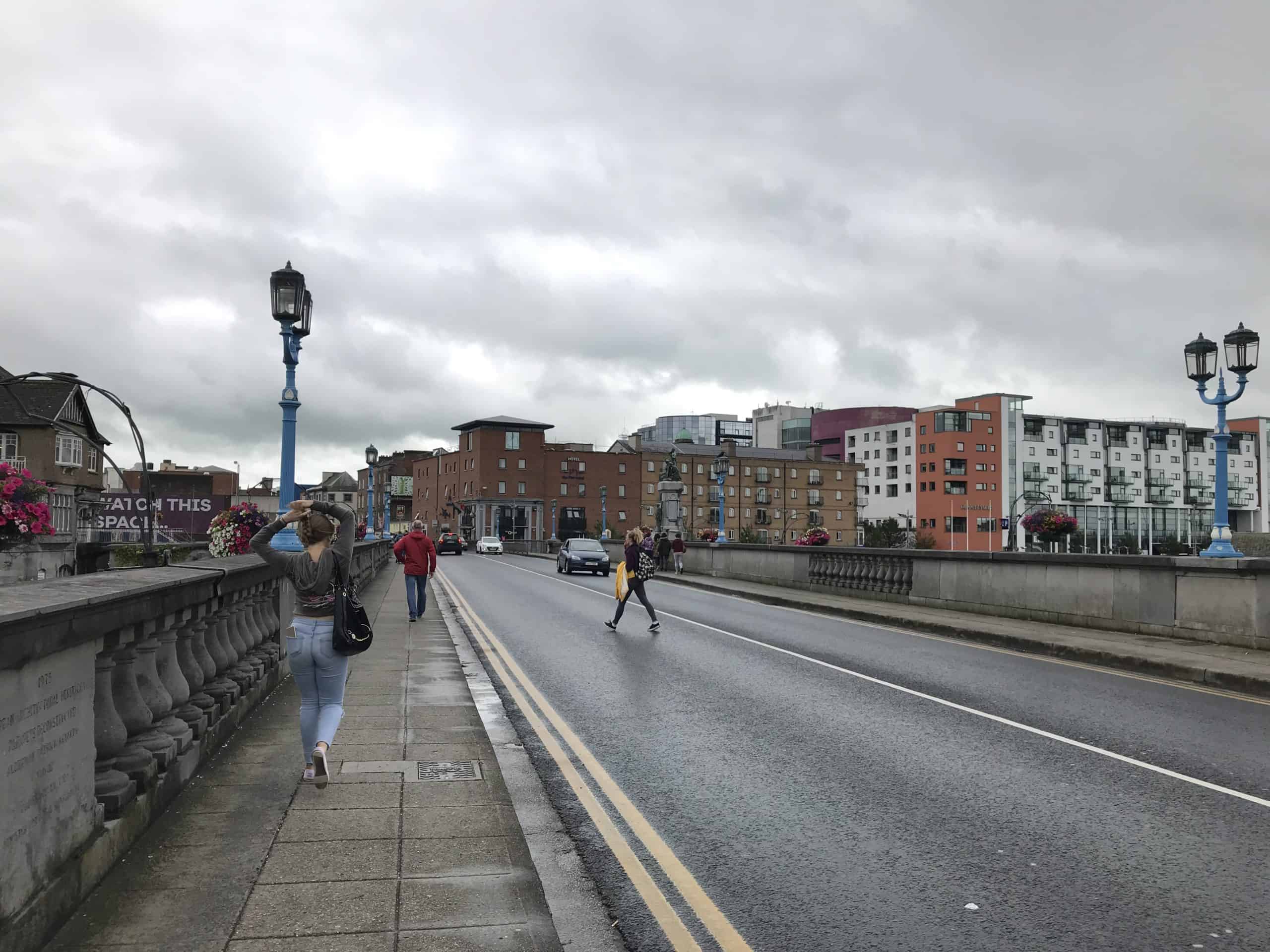 In Limerick, Ireland. A bridge with a road and sideway. A few local people are walking around. Behind the bridge are red, brown, white buildings. A dark cloudy sky.