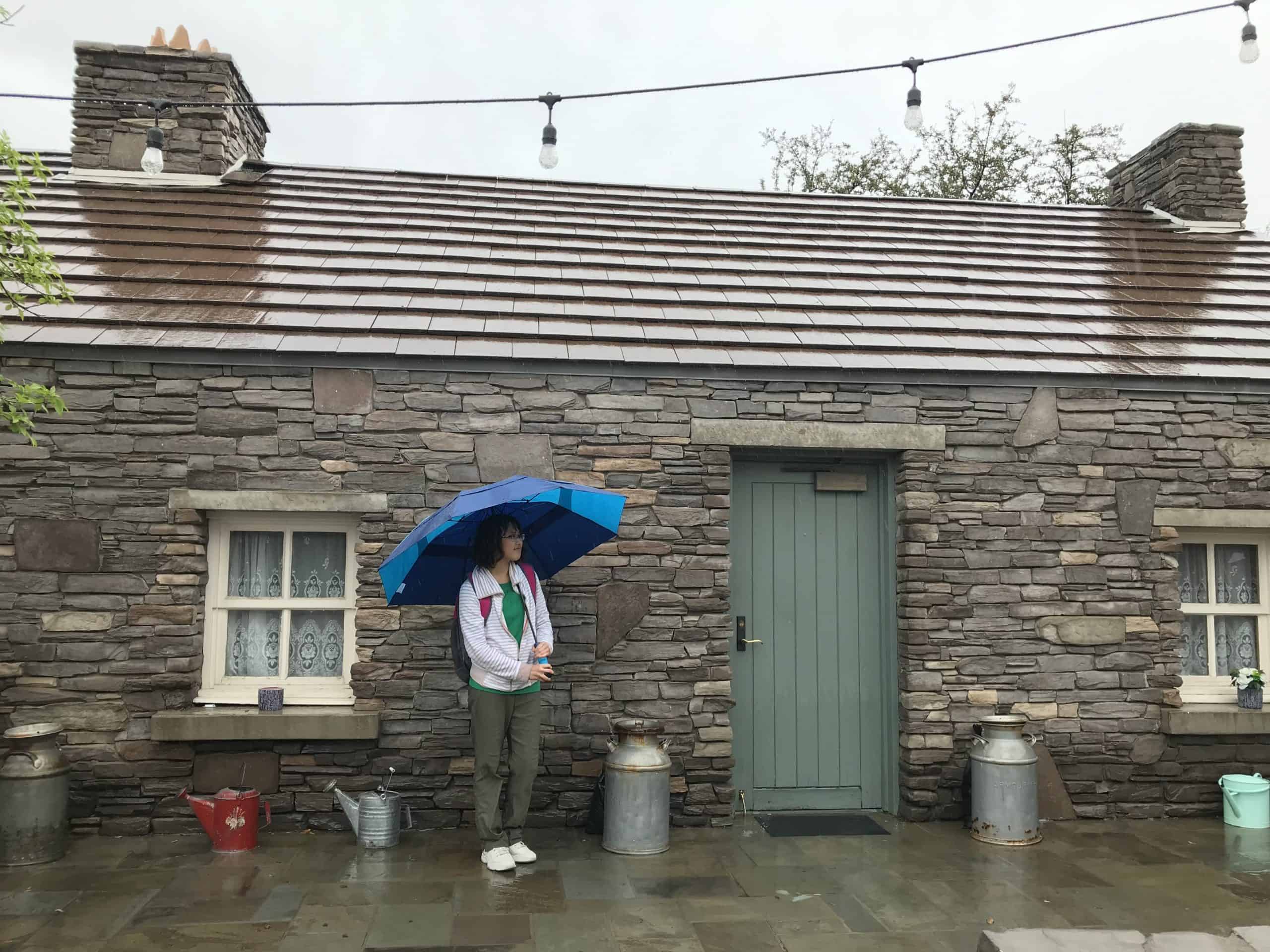 In Phoenix, Arizona, United States's Irish Cultural Center. Meggie is standing underneath a blue umbrella as it's raining. Behind her is a replica traditional Irish stone cottage.