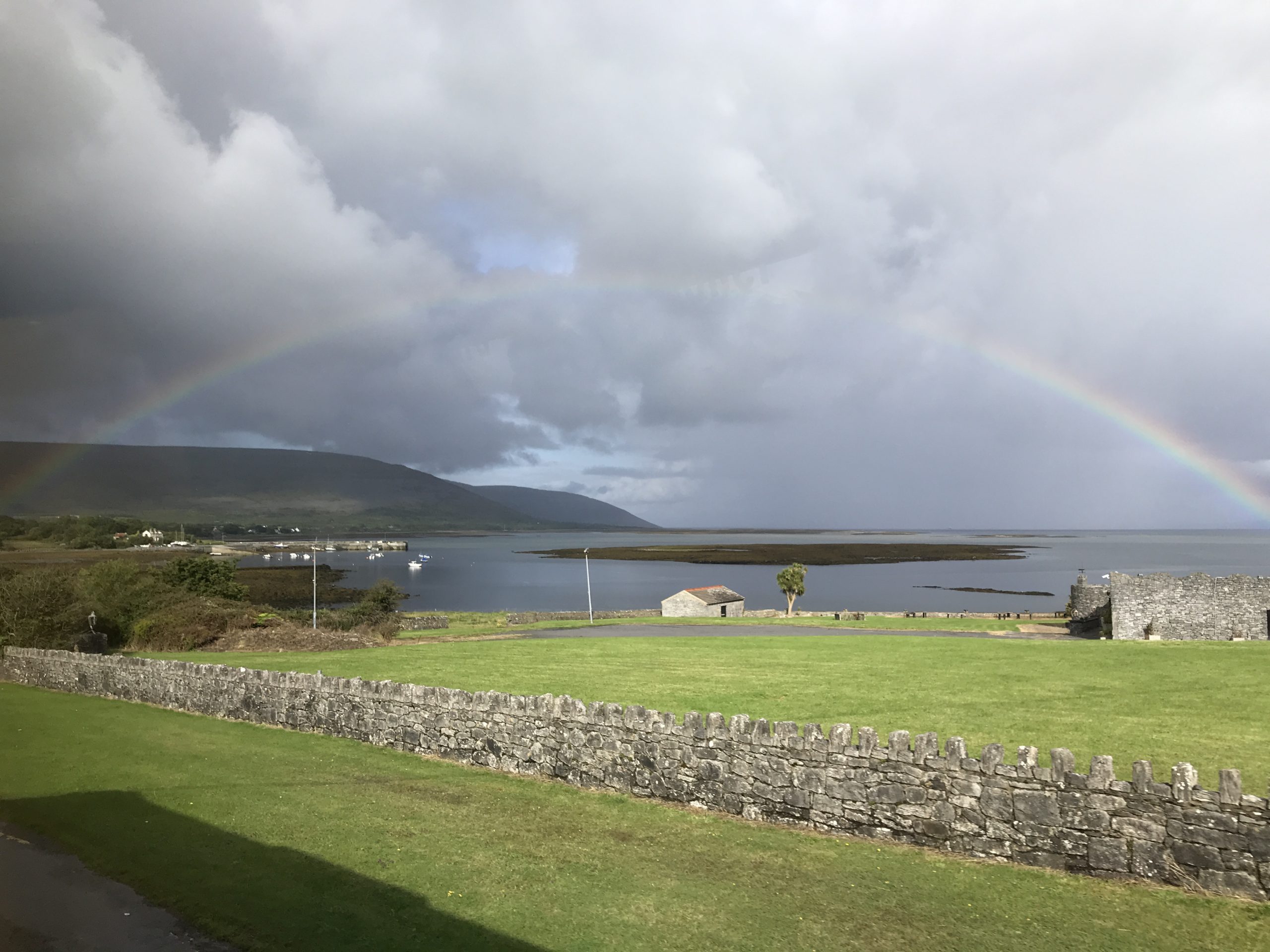 Green grass next to a lake with mountains in the background. Partly cloudy. A large rainbow spans over the water. Along the Wild Atlantic Way between Galway and Doolin, Ireland.