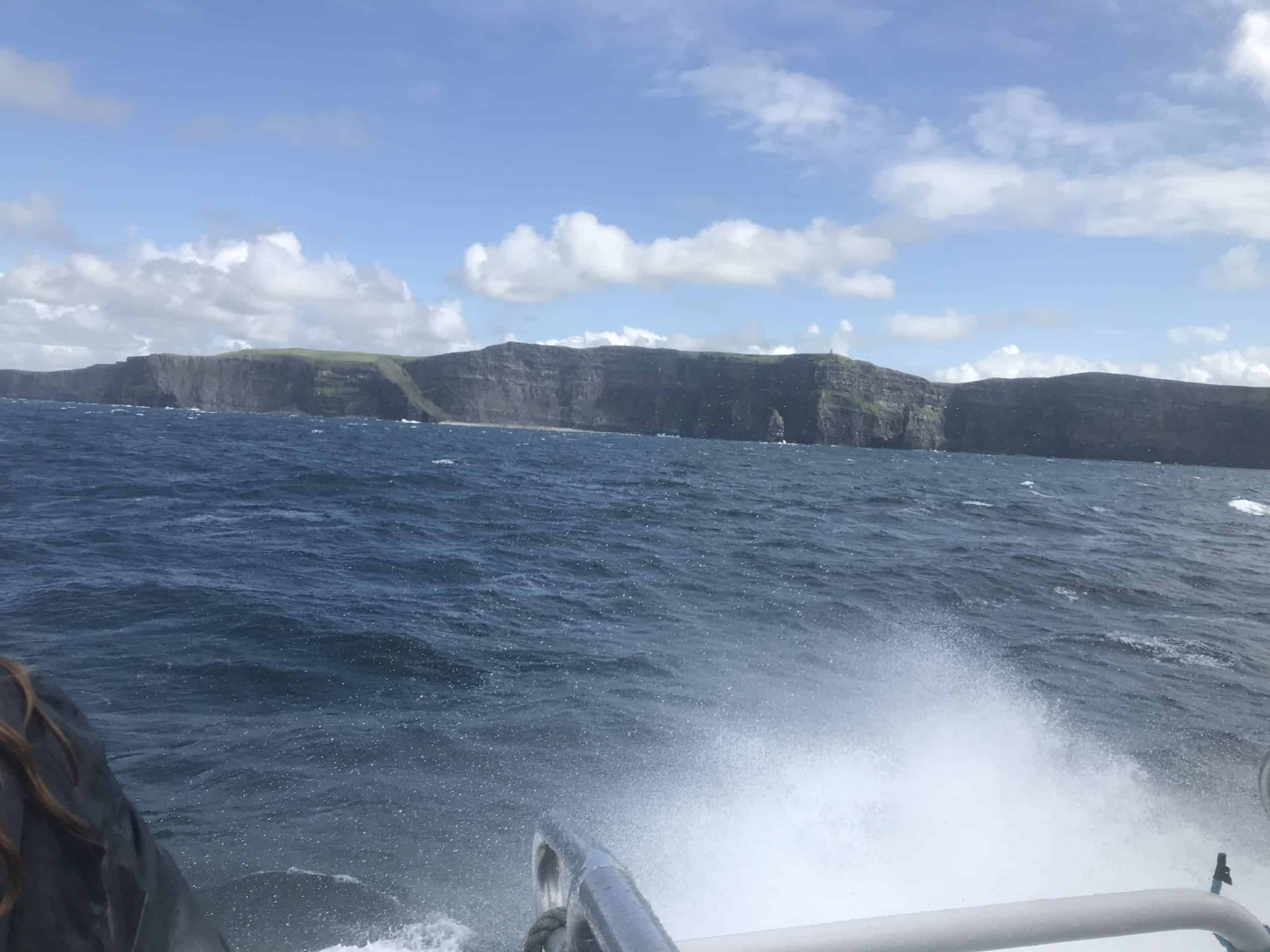Blue sunny sky with some clouds. Cliffs of Moher in the distance as I'm riding a boat. A white wave makes a big splash. Choppy rolling waves of the blue Atlantic Ocean. Nearby Doolin along the Wild Atlantic Way, Ireland.