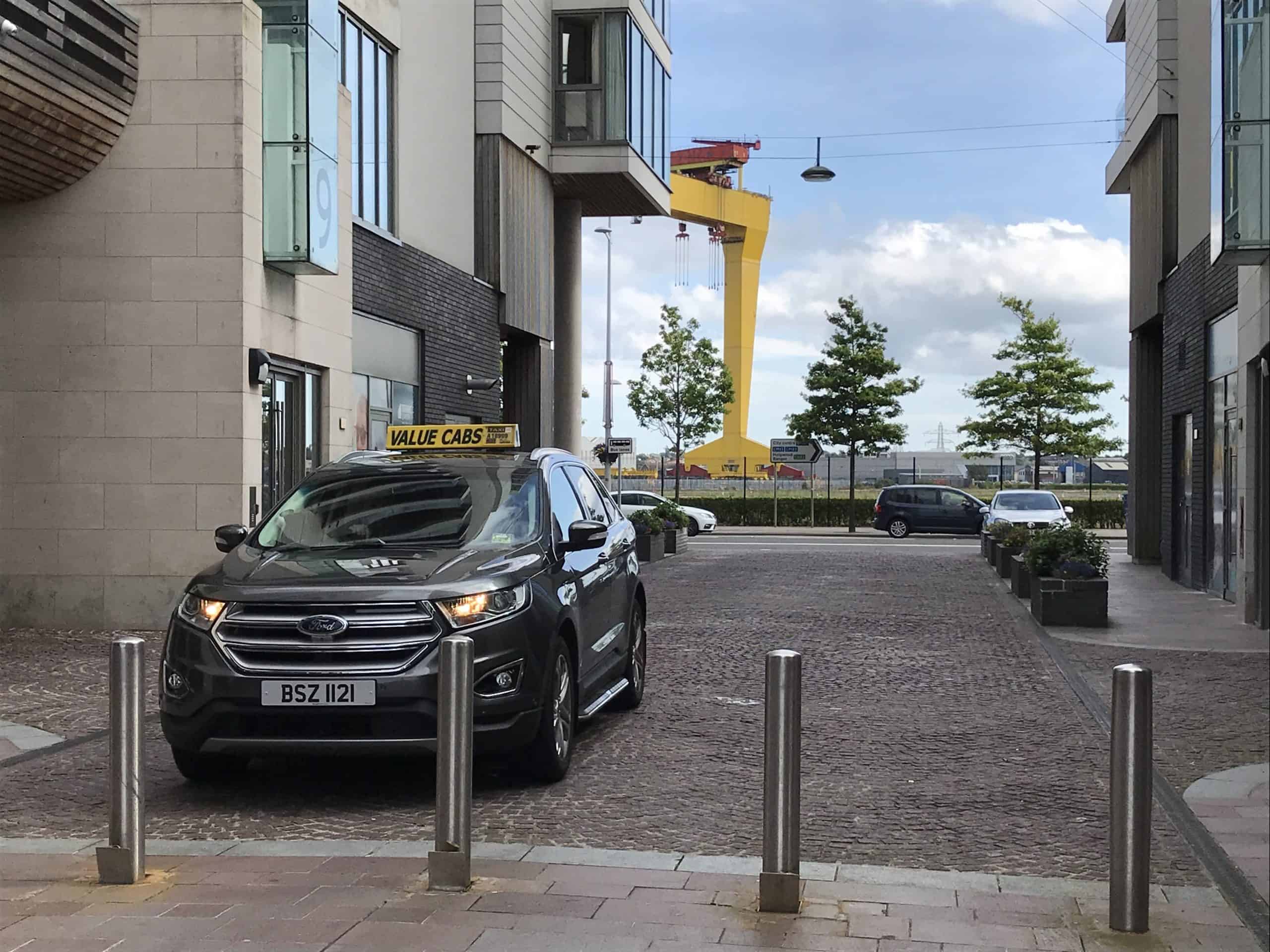 A dark gray SUV as a taxi cab. Parked in a small alley. White and gray apartment buildings along the alley. Behind is the blue sunny partly cloudy skyline with the iconic yellow Samson and Goliath shipbuilding gantry cranes in Belfast, Northern Ireland.