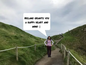 Meggie is standing proudly between two green hills on a paved walkway under a cloudy sky. In the comic speech bubble, she says: ireland grants you a happy heart and mind. Photo taken at Carrick-a-Rede rope bridge along the Causeway Coastal Route in Northern Ireland.