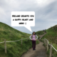 Meggie is standing proudly between two green hills on a paved walkway under a cloudy sky. In the comic speech bubble, she says: ireland grants you a happy heart and mind. Photo taken at Carrick-a-Rede rope bridge along the Causeway Coastal Route in Northern Ireland.