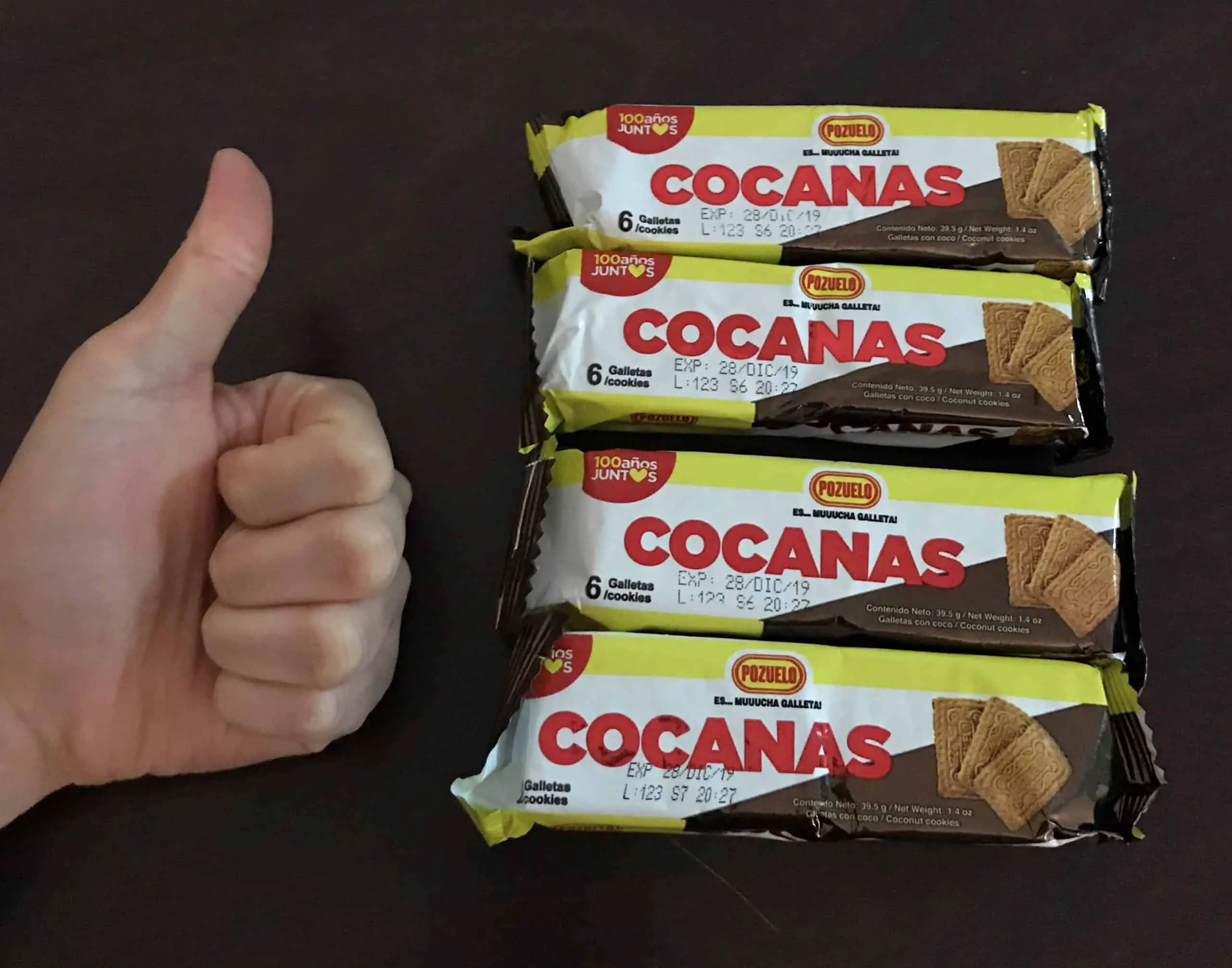 On a wood table are four Costa Rican Coconas coconut cracker snacks in their unopened wrappers. Meggie's hand, in a thumbs up, is next to the crackers.