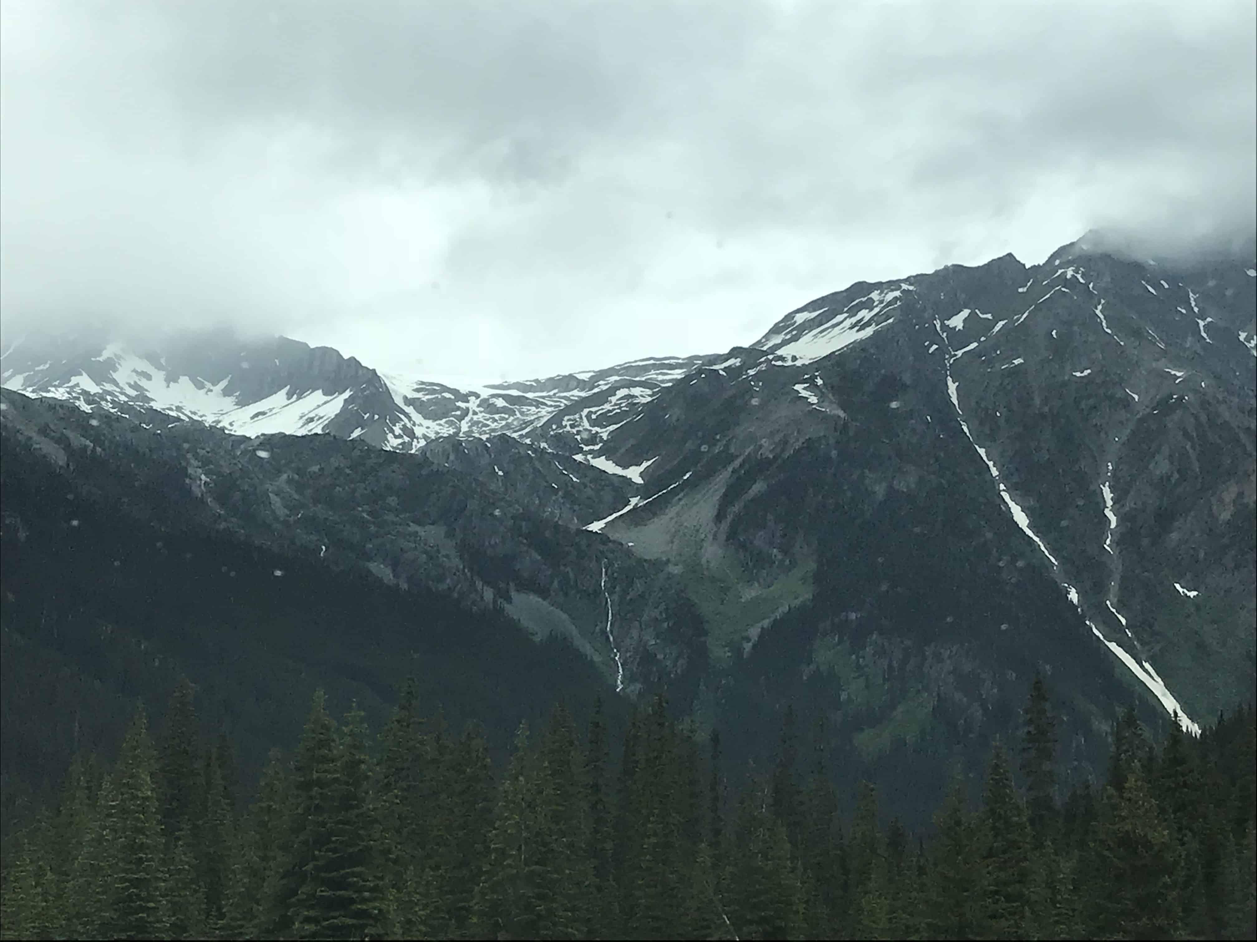 A tall, dark gray mountain range covered with snow overlooks a pine forest. A dark cloudy overcast. In the Canadian Rockies along the Trans-Canada Highway between Vancouver, British Columbia and Calgary, Alberta.