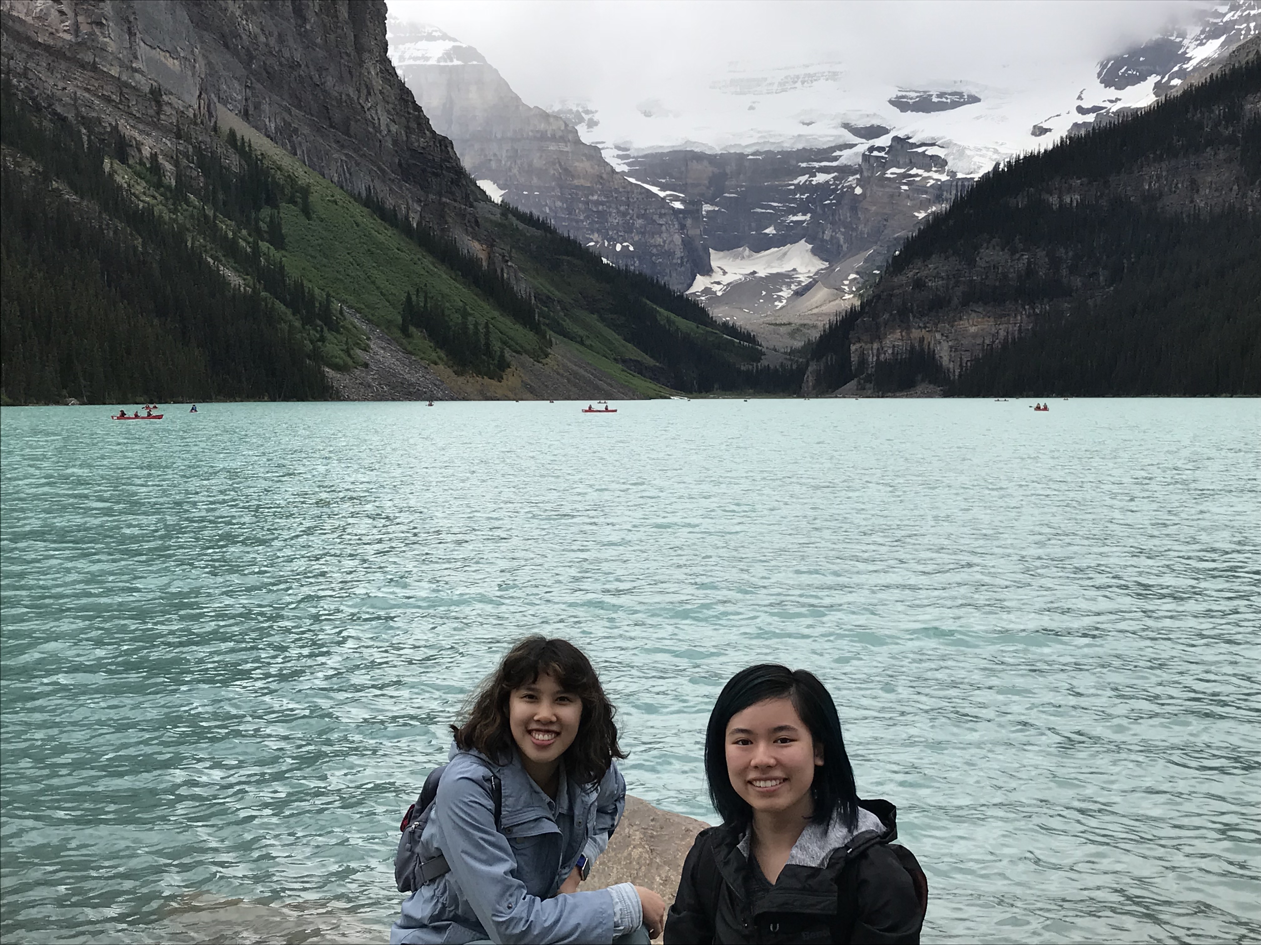 At Lake Louise in Banff National Park, Alberta, Canada. The turquoise lake has people rowing in canoes. Behind the lake is a tall, dark gray mountain range covered with snow, and a dark green pine forest covers the lower parts. Meggie, in her blue jacket, and Val, in her black jacket, are smiling in front of the whole scene.