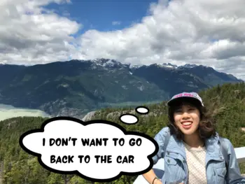 Meggie is standing on a balcony overlooking Howe Sound, pine forests, a turquoise sea, and mountains. Meggie is smiling, wearing a ballcap, a light blue denim jacket, and a white tank-top with black polka dots. The brilliant blue sky gas a few large clouds. Sea to Sky Gondola nearby Squamish, British Columbia, Canada. A thought bubble next to Meggie has the words, "I don't want to go back to the car"