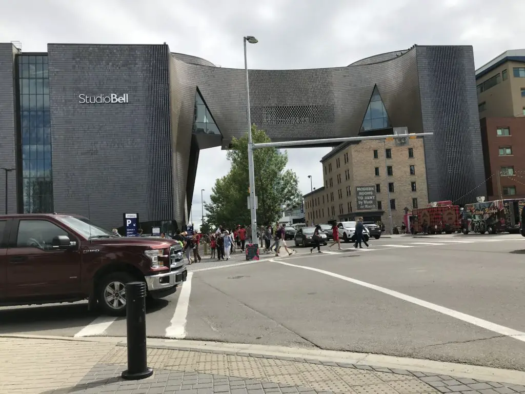 The exterior of downtown Calgary, Alberta, Canada's Studio Bell's National Music Centre. The building hangs over the road. The building has dark grey, shiny bricks and several futuristic-looking window panes.