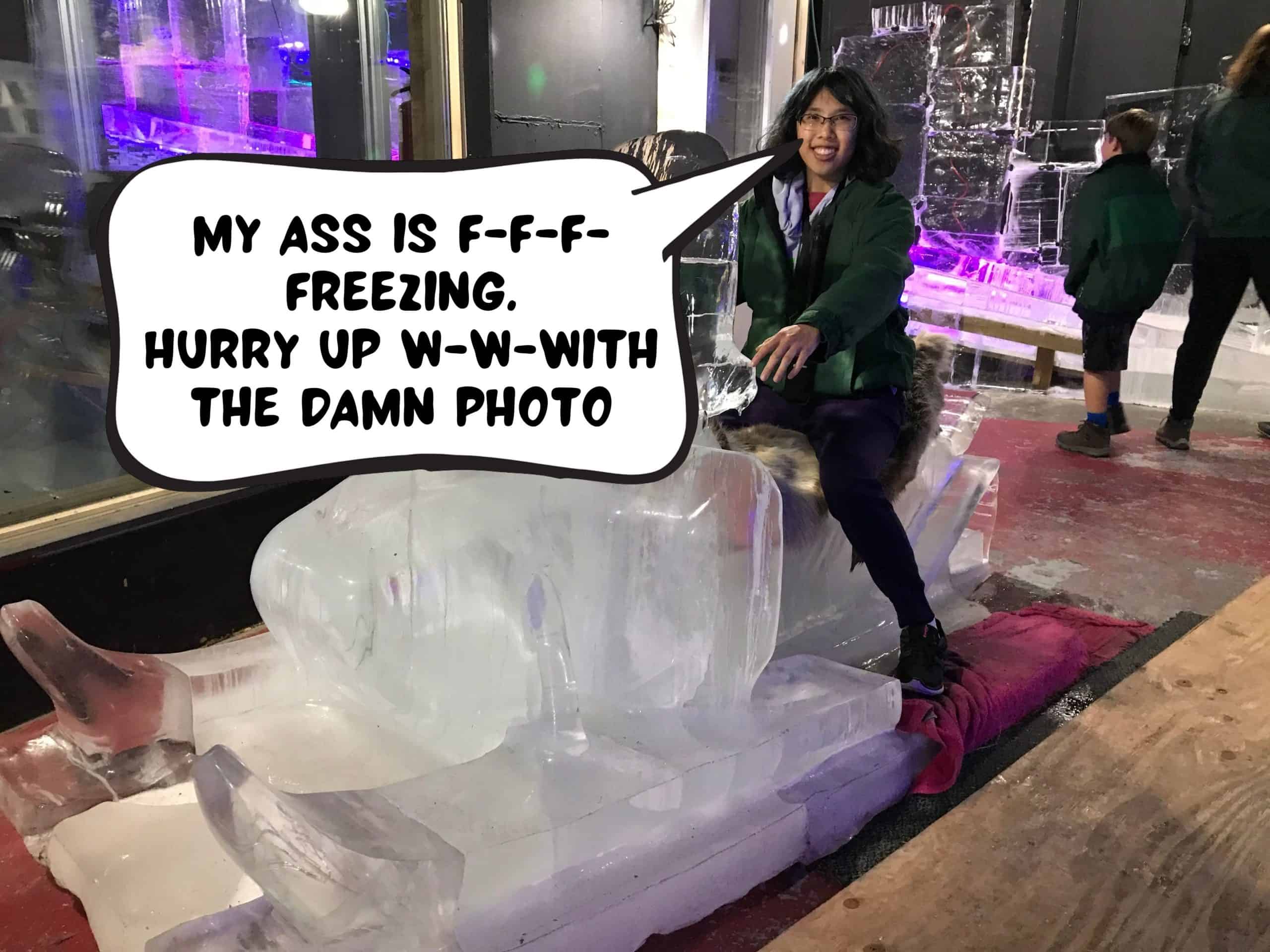 Meggie is sitting on a block of ice shaped into a snowmobile. She is wearing a green jacket. She is smiling. The comic speech box in front of her says, "My ass is f-f-f-freezing. Hurry up w-w-with the damn photo." At the Ice Museum in Downtown Fairbanks, Alaska, United States