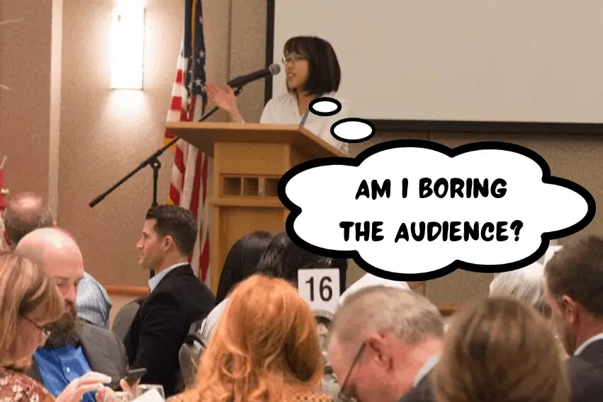 Meggie is standing on a stage giving a speech to a large business audience. She has her hand out and open as she is speaking. She is wearing a white business suit.