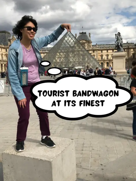 Meggie, with her blue jacket and purple shirt and pants, is standing on top of a column in front of the Louvre Art Museum in Paris, France. She is pinching her fingers as if she is grapping the top of the Louvre glass pyramid. A comic thought bubble says, "Tourist bandwagon at its finest."