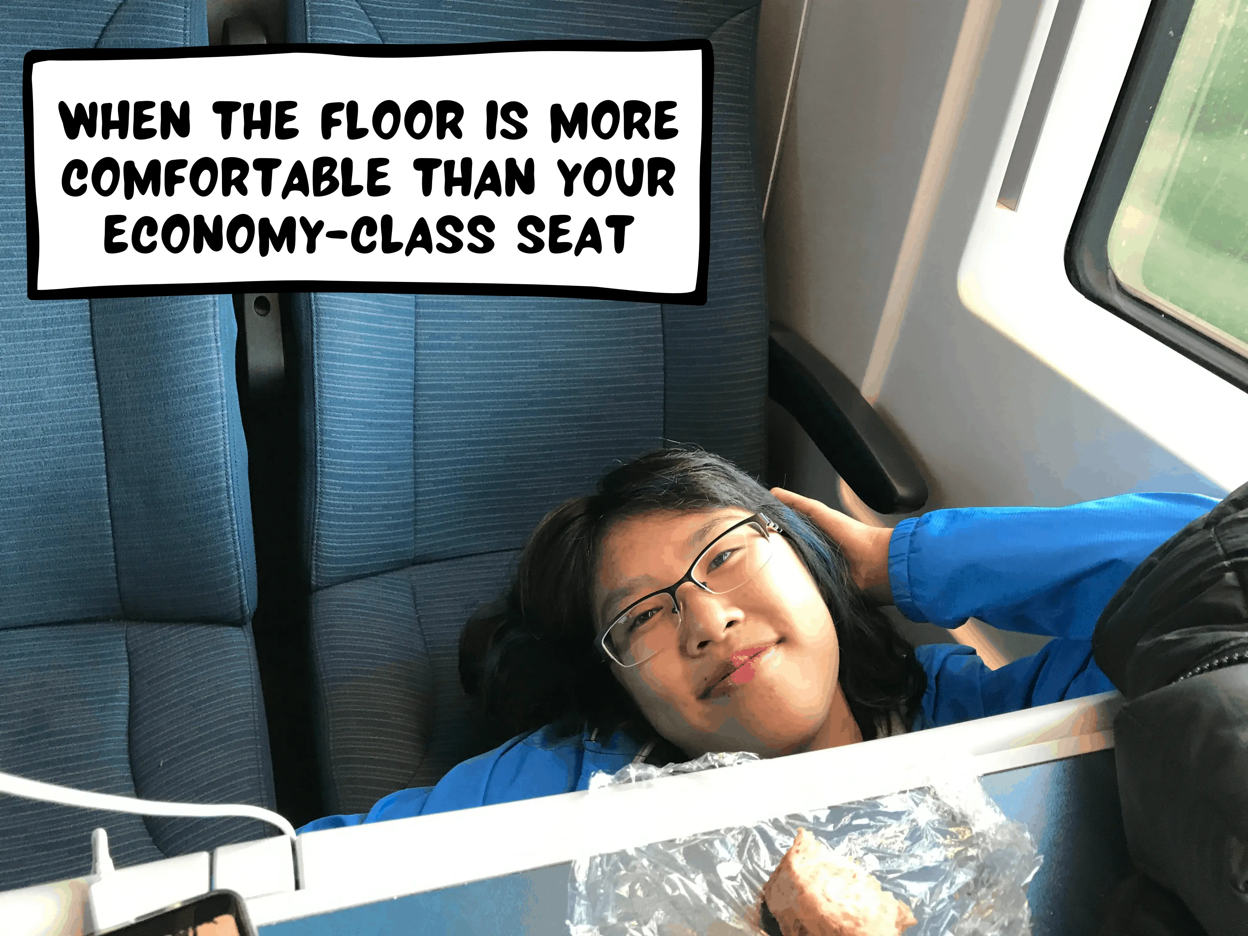 Meggie in her blue jacket is underneath the table in the Eurostar commuter train between Paris and London. She is smiling in a cheeky way. The comic text box says: "when the floor is more comfortable than your economy-class seat"
