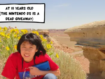 Meggie as an 11 year old with a red t-shirt with the St Louis Missouri Gateway Arch is sitting in front of a desert in Colorado or Utah, United States. The desert scene is brown and rocky. She is holding a Nintendo DSi XL. Yellow wildflowers are growing behind her.