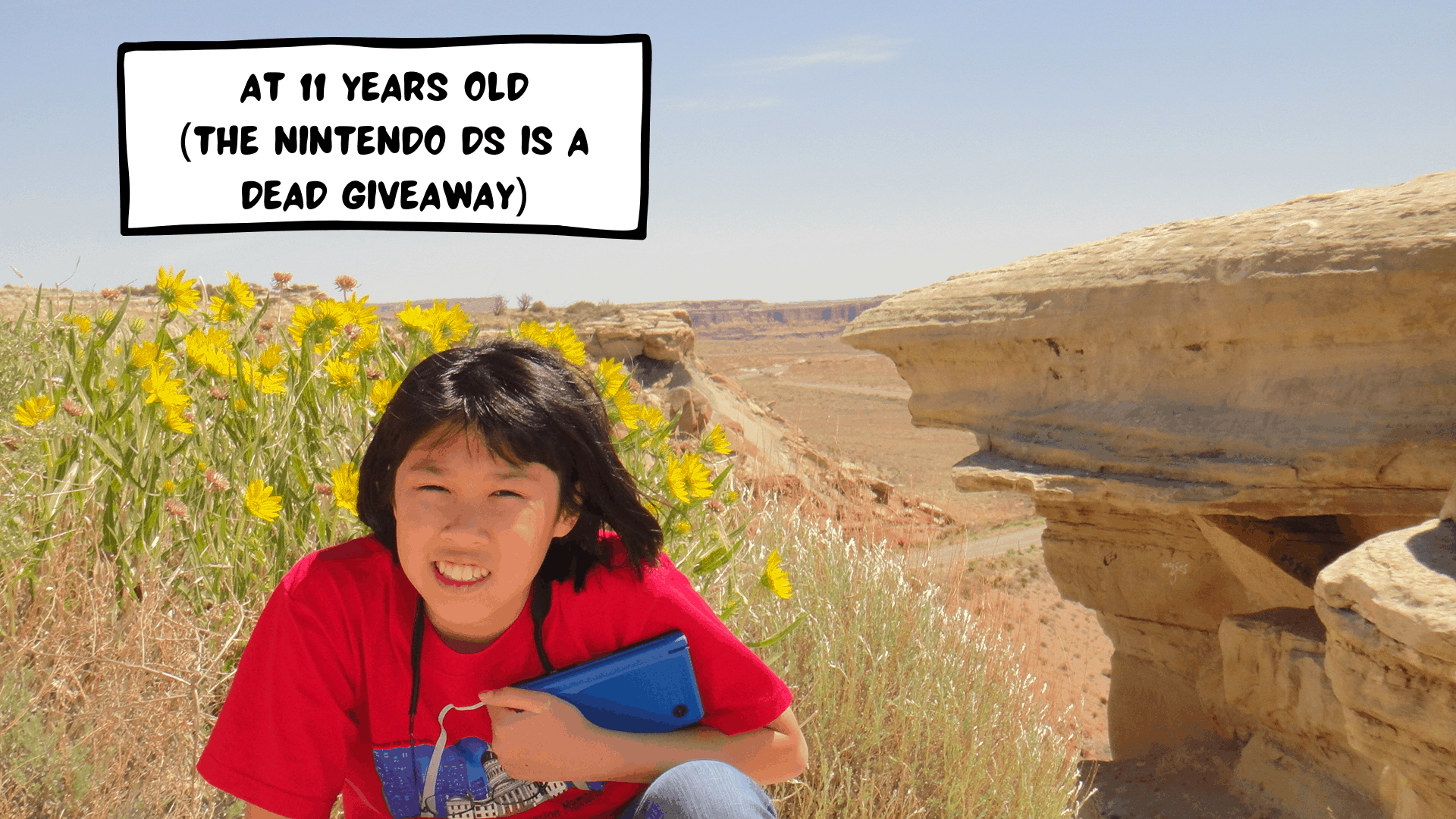 Meggie as an 11 year old with a red t-shirt with the St Louis Missouri Gateway Arch is sitting in front of a desert in Colorado or Utah, United States. The desert scene is brown and rocky. She is holding a Nintendo DSi XL. Yellow wildflowers are growing behind her.