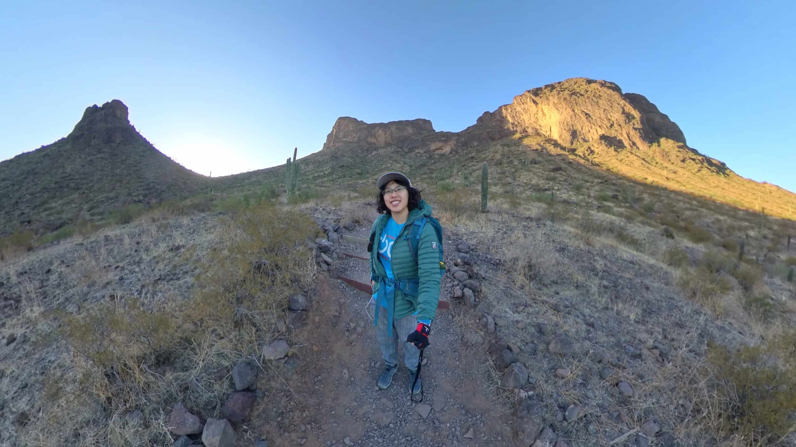 Meggie is wear a blue shirt that says "OCD" and has on red and black gloves. She is wearing a blue backpack and ballcap. She is at the base of the mountains. Behind her is the vast brown desert and cactus and a clear blue sky. She is in Picacho Peak State Park between Tucson and Phoenix, Arizona, United States.