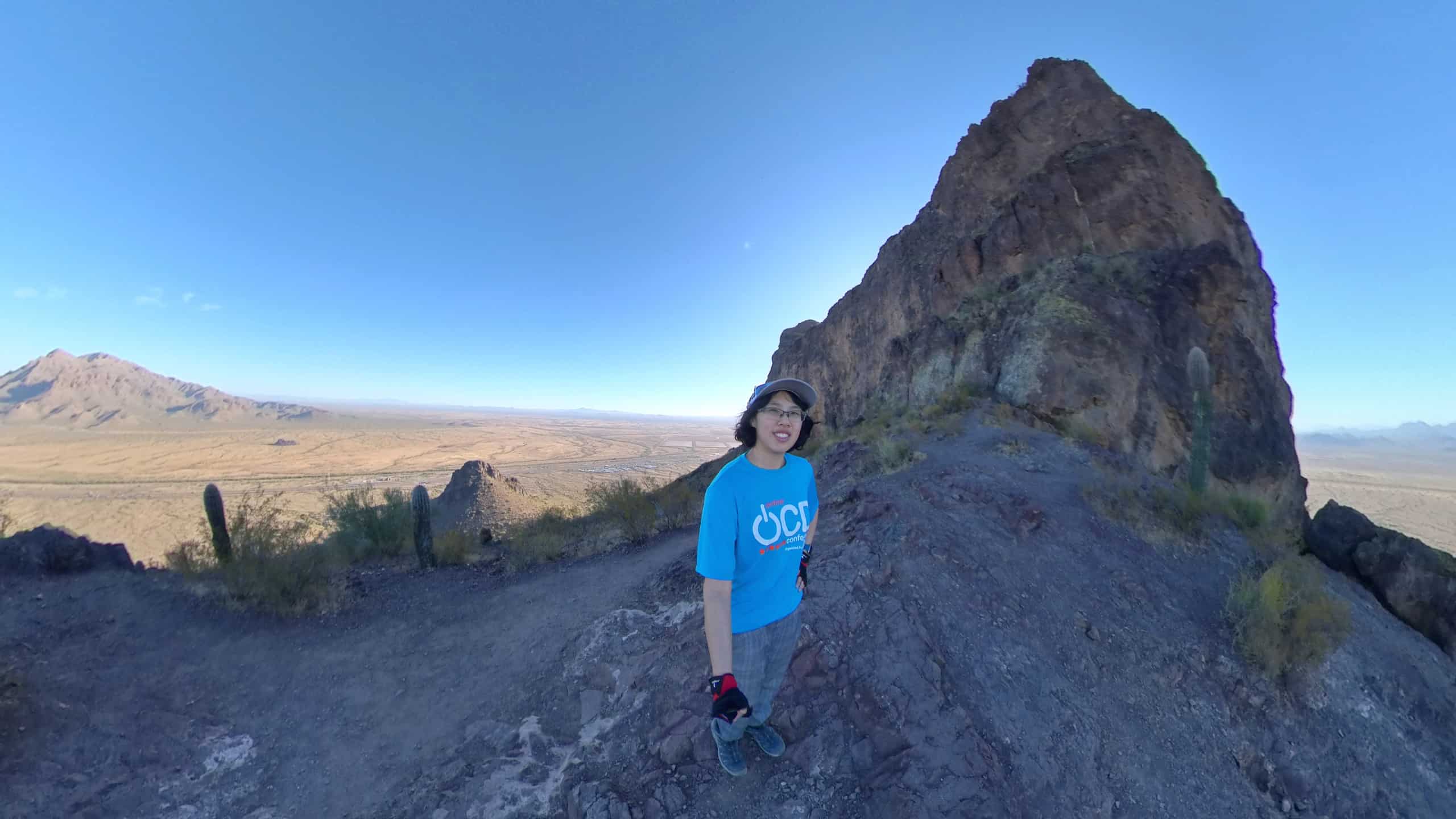 Meggie is wear a blue shirt that says "OCD" and has on red and black gloves. She is wearing a blue backpack and ballcap. She is in the shadows of a large brown mountain. She is on the saddle between the mountains. Behind her and the mountain is the vast brown desert and cactus and a clear blue sky. She is in Picacho Peak State Park between Tucson and Phoenix, Arizona, United States.