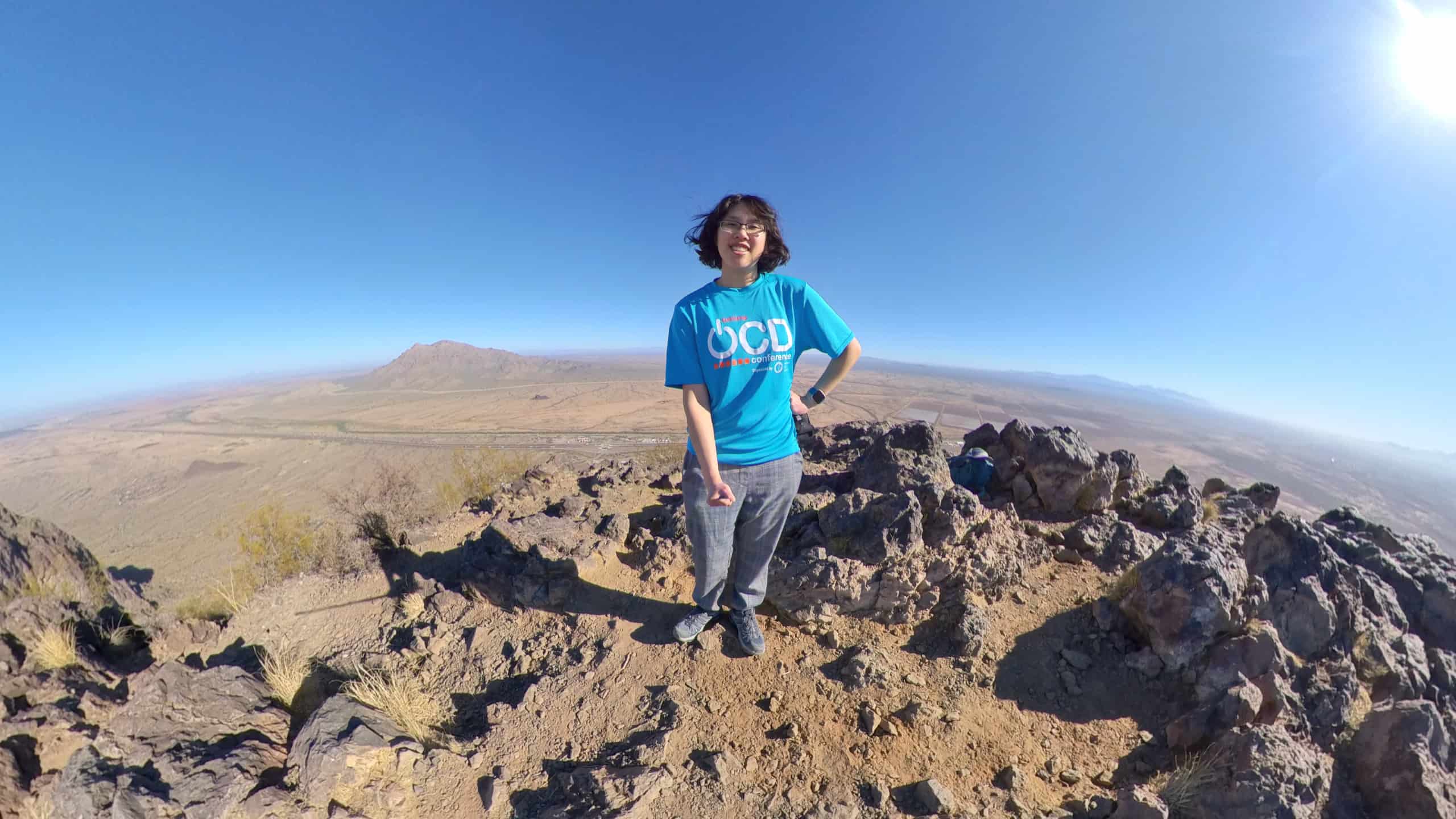 Meggie is wear a blue shirt that says "OCD" and has on red and black gloves. She is wearing a blue backpack and ballcap. She is standing on the mountain summit of Picacho Peak. Jagged rocks are next to her. Behind her is the vast brown desert, interstate 10, the railroad, and a clear blue sky. She is in Picacho Peak State Park between Tucson and Phoenix, Arizona, United States.