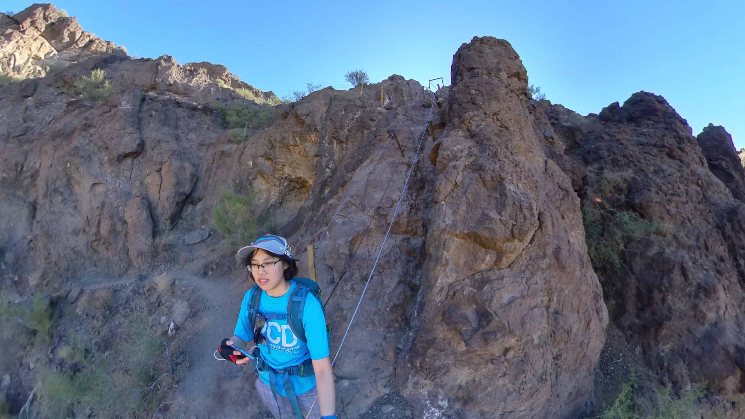 Meggie is wear a blue shirt that says "OCD" and has on red and black gloves. She is wearing a blue backpack and ballcap. She is in the shadows of a large brown mountain. Behind her are the cables scaling up the steep mountainside. She is in Picacho Peak State Park between Tucson and Phoenix, Arizona, United States.