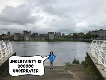 Meggie with her blue jacket is standing proudly at a boat ramp leading down to the water of the River Shannon in downtown Limerick, Republic of Ireland. The sky is gloomy, dark, and full of clouds. Behind her and the river is the grey bricked King John's Castle. A comic text bubble from Meggie says, "Uncertainty is so overrated"