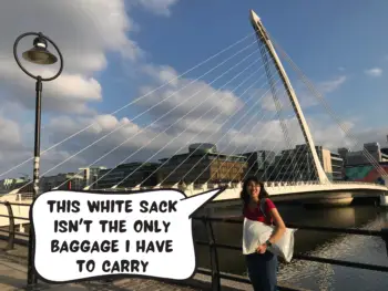 Meggie is wearing a black and red striped shirt as she is holding a white sack. She is standing in front of the Samuel Beckett white harp bridge in Dublin, Republic of Ireland. The bridge goes over the River Liffey. A blue sky with some clouds hangs over. Meggie, in a comic text box, says "this white sack isn't the only baggage i have to carry"