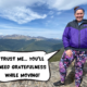 Allie from the Hoppy Passport is standing on a gray mountainside overlooking the forest and a mountain range. She is wearing a black jacket and floral pants. In a text comic bubble, she says, "Trust me... you'll need gratefulness while moving!"