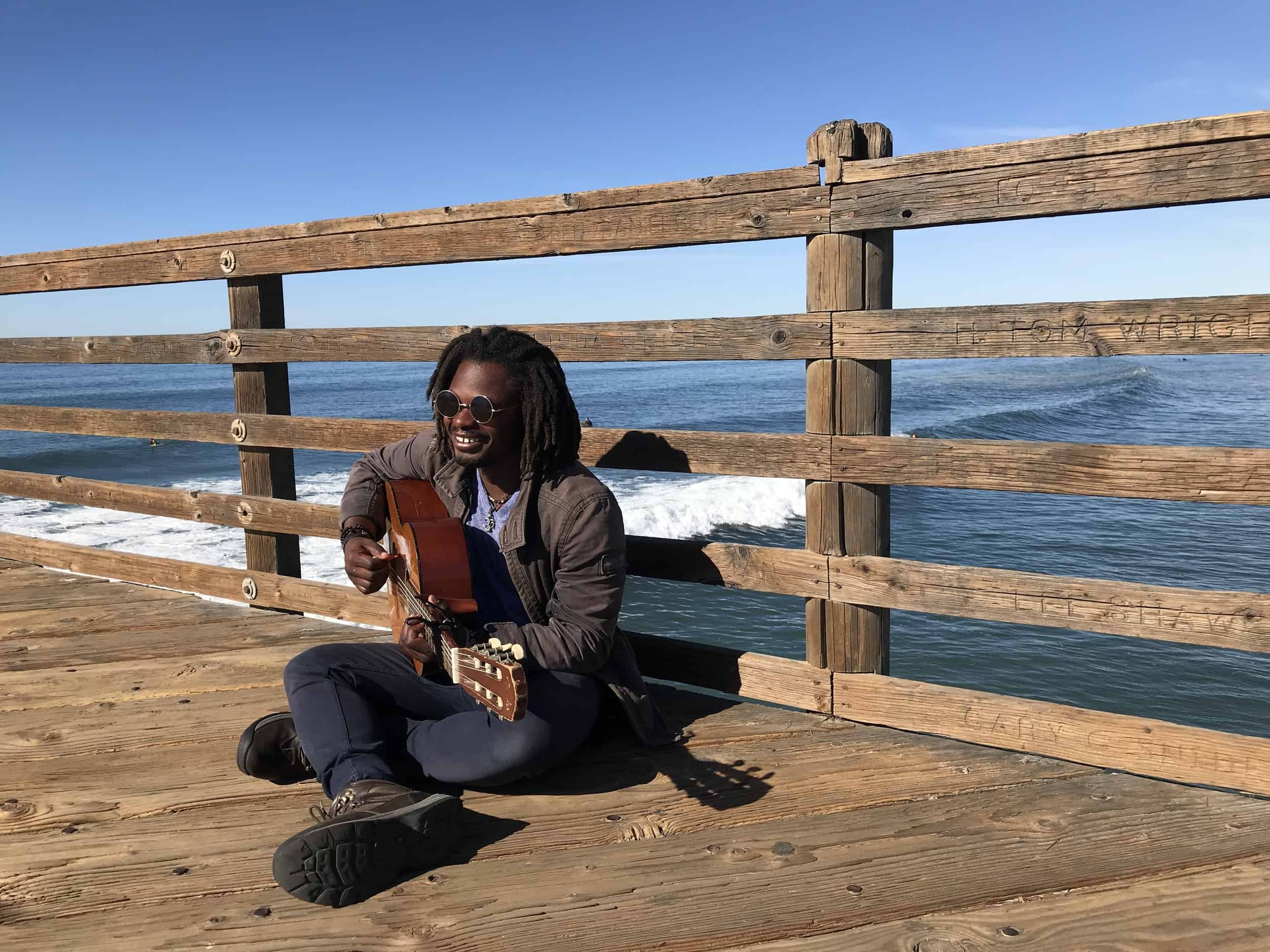 The kowboy rasta musican plays his acoustic guitar while sitting on the pier's floor. he is leaning against the fence of the pier. Beyond the pier is the Pacific Ocean and a long ocean wave. Dark blue waters. Oceanside Pier in downtown Oceanside, California, United States of America