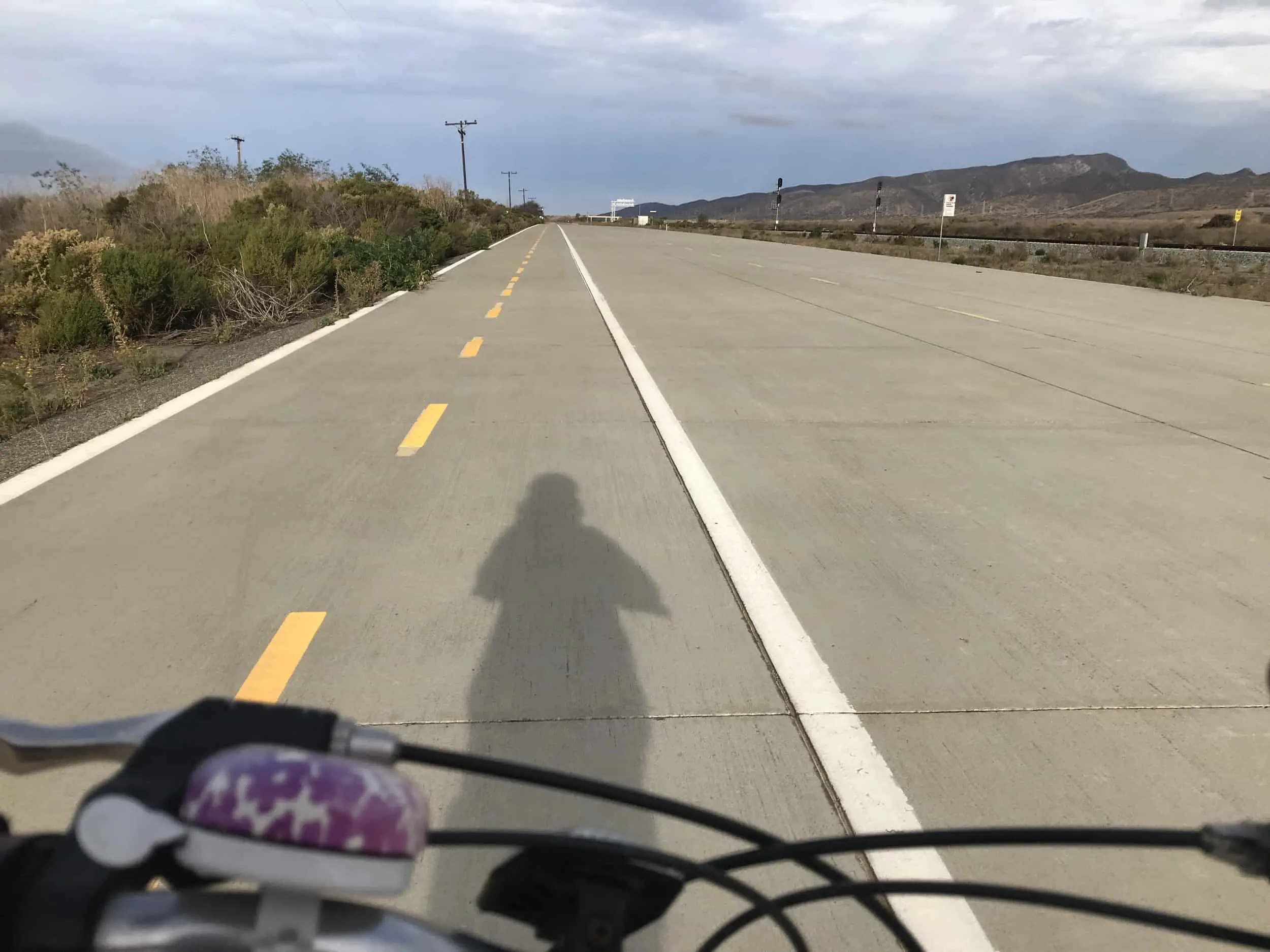 a paved bike lane running next to the Pacific Ocean in a desert brush landscape. brown bushes and mountains and grass. cloudy sky. Meggie's shadow can be seen from her bike. Her bike's handlebars are shown. Las Pulgas/Old Pacific Highway Trail nearby Oceanside and San Clemente, California, United States of America