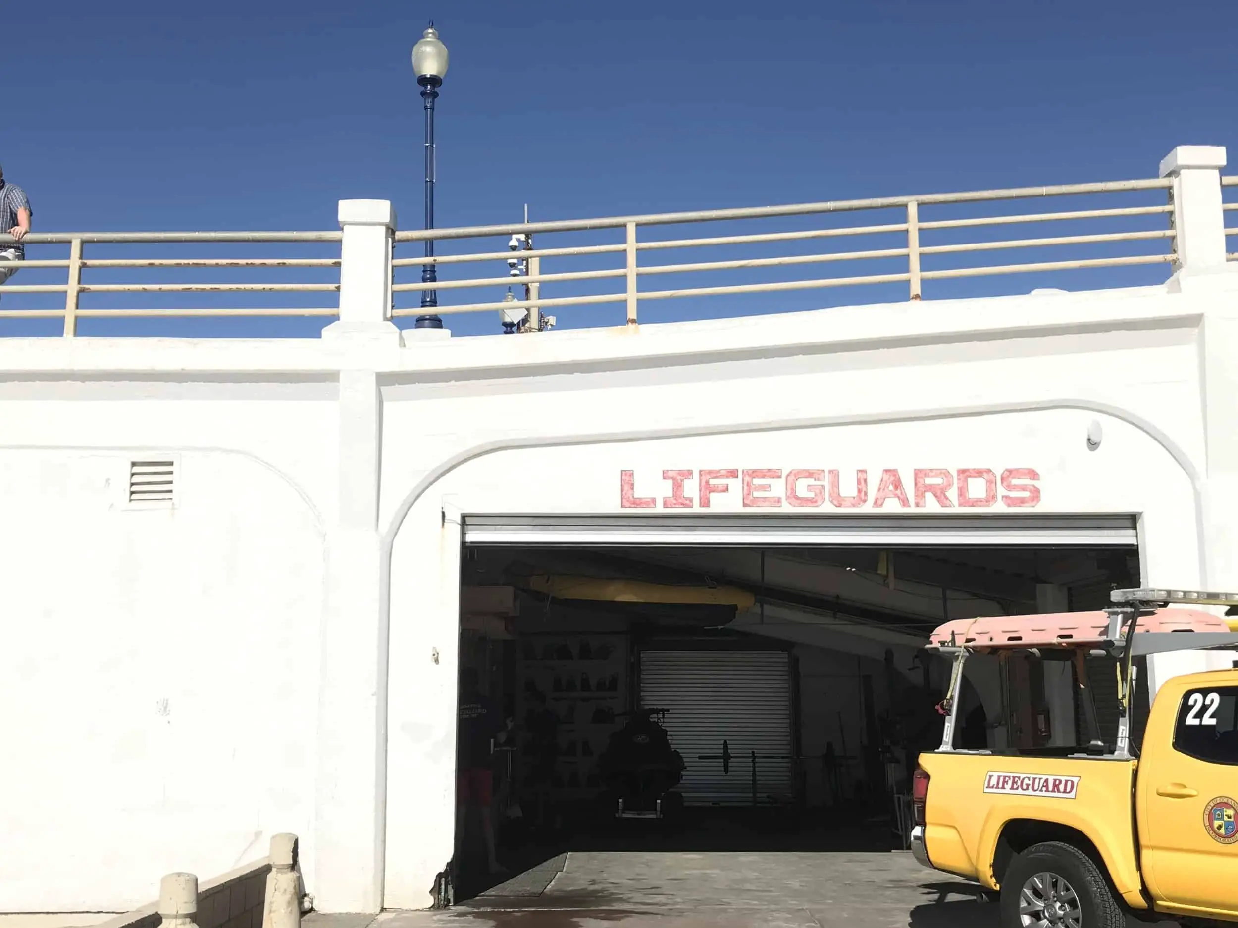 A clear blue sky over the white painted pier. Underneath the pier is a lifeguard headquarters with gym and swim equipment. A yellow truck with surfboards is parked outside the garage. the word "lifeguards" is painted in red above the garage. Oceanside Pier in downtown Oceanside, California, United States of America