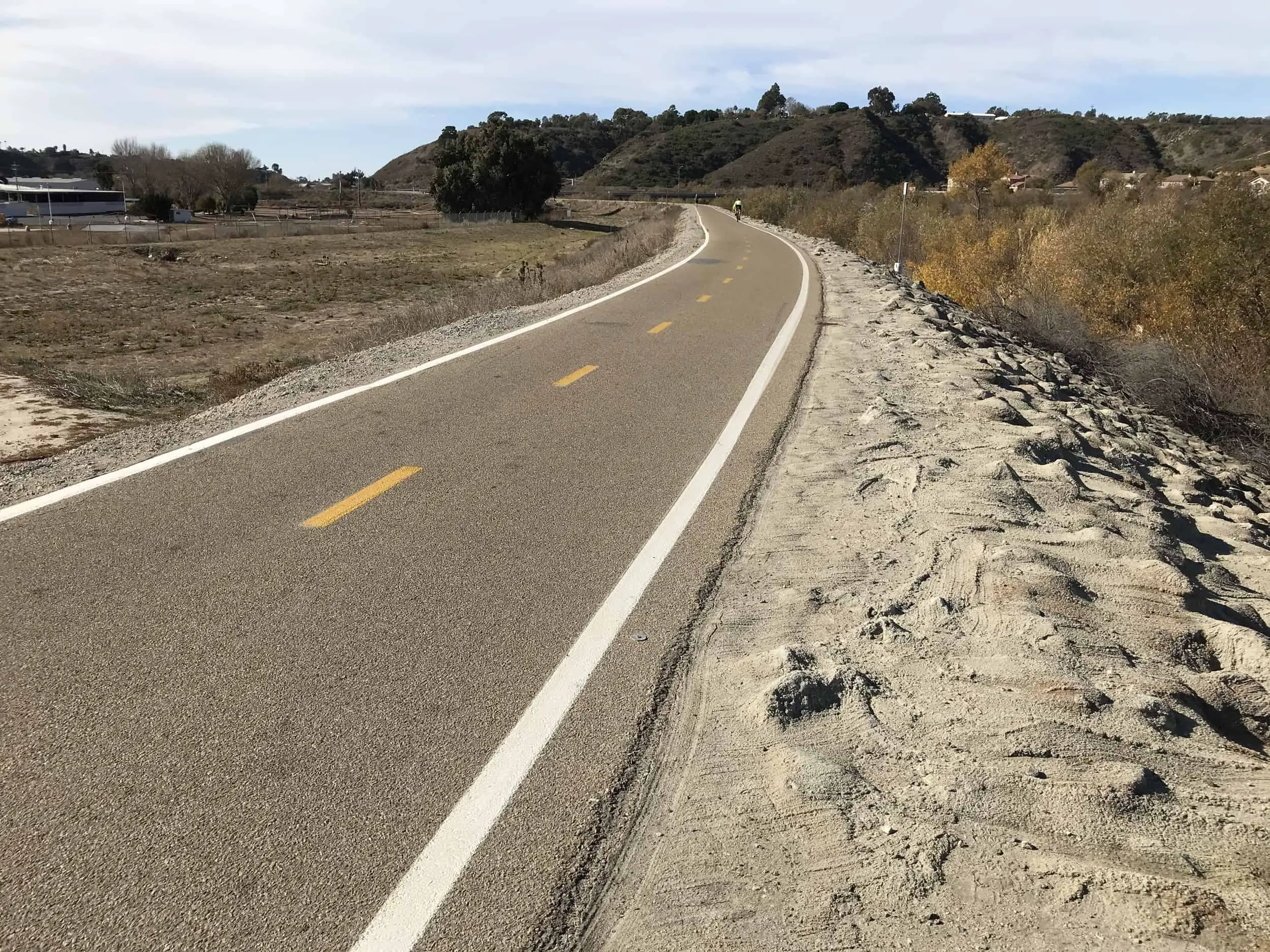 a paved bike lane running through a desert brush landscape. tan bushes and grass and hills. partly cloudy sky. San Luis Rey River Trail in Oceanside, California, United States of America
