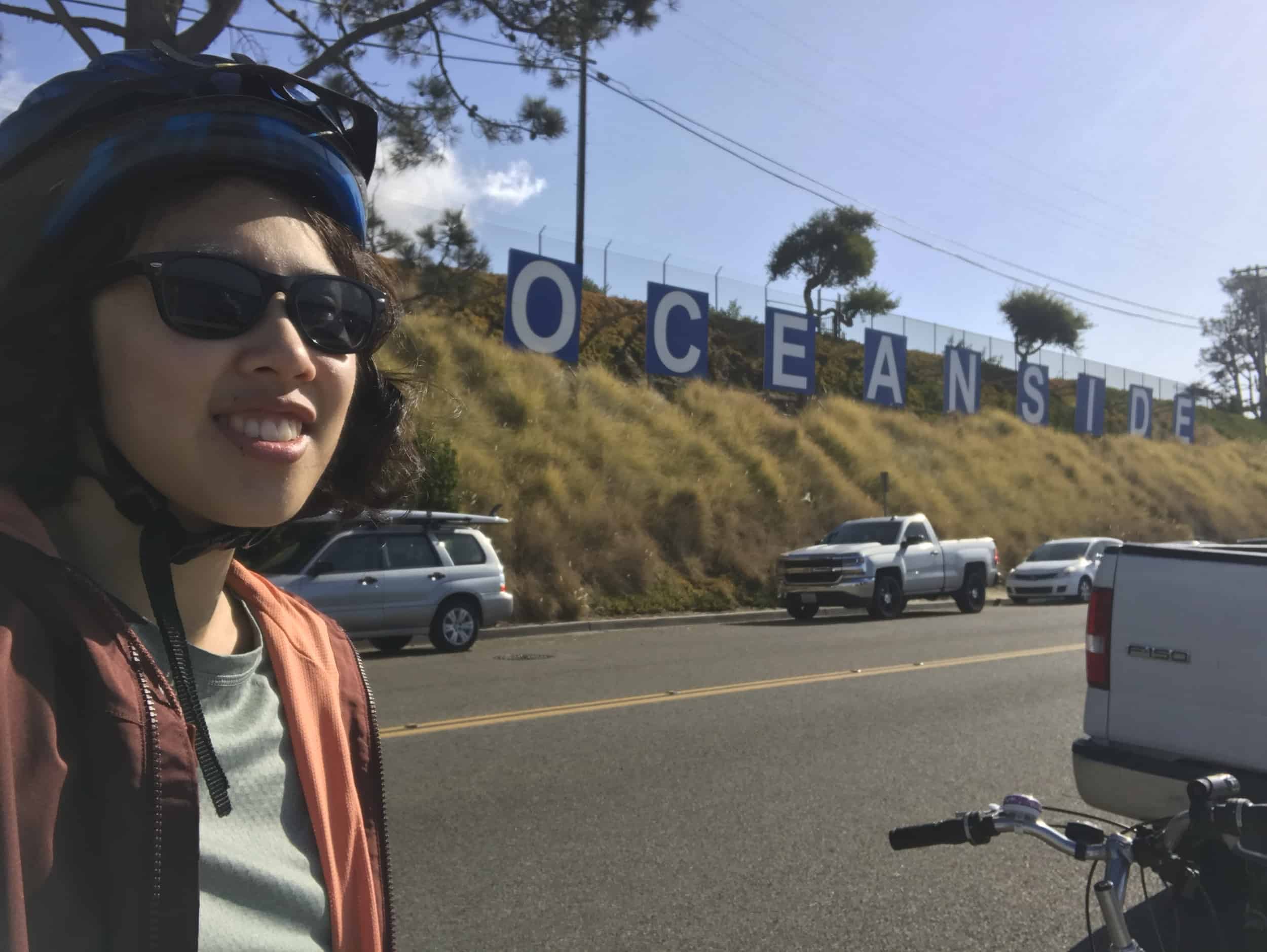 Meggie is wearing sunglasses, a maroon jacket, and a blue bike helmet. She is standing in front of a series of blue and white signs spelling "Oceanside" along the desert grassy hillside. Harbor Beach in Oceanside, California, United States of America