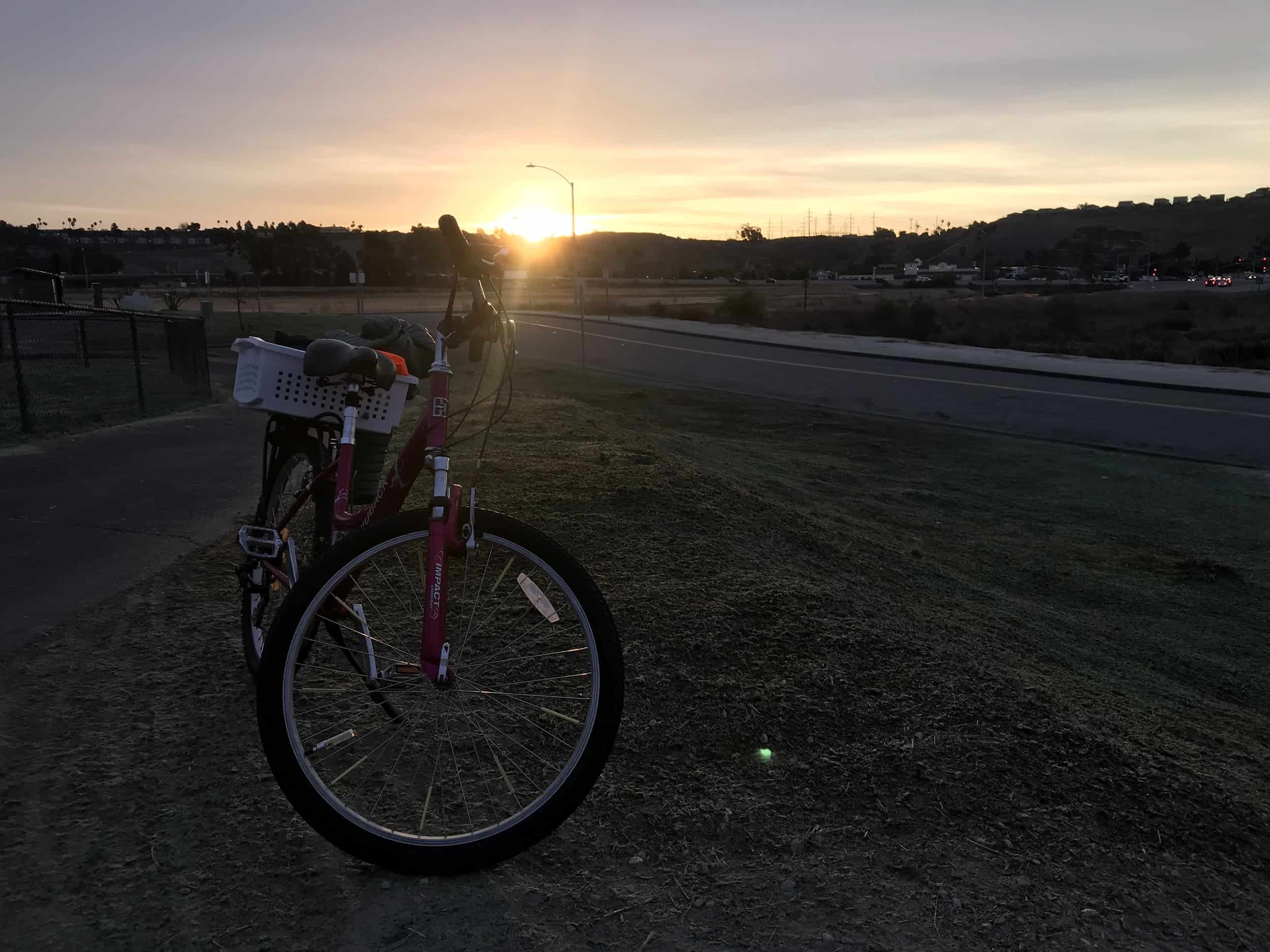 Meggie's bike is on top of a grassy hill. The sun is just rising so everything is dark except the yellow and orange sky of a new day amongst the clouds.  Prince Park/Alex Road Skatepark on the San Luis Rey River Trail, Oceanside, California, United States of America