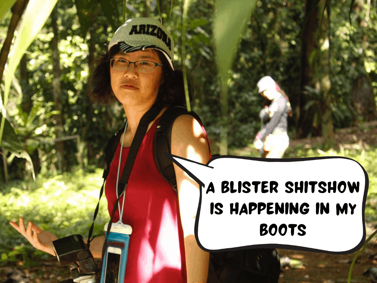 Meggie is hiking through the jungle rain forest with a friend behind her. Meggie is underneath a canopy of leaves. Green leaves and trees and plants all over the place. A brown foot trail runs through. Meggie has her arms open and she has a grimace on her face. She says in a comic text bubble, "A blister shitshow is happening in my boots." The La Selva Biological Station in Puerto Viejo de Sarapiquí in the Heredia Province of Costa Rica, Central America.
