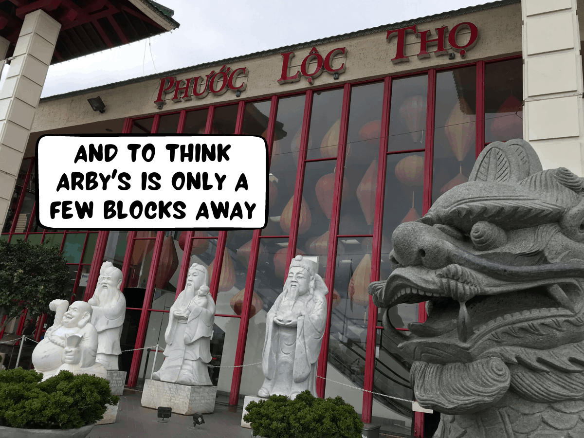 Statues of a lion and powerful human figures stand in front of a famous Vietnamese shopping building called Phuoc Loc Tho, with its red decor and lettering, in Asian Garden Mall, Bolsa Avenue, Westminster, Little Saigon, Orange County, California, United States of America. A comic text bubble says, "And to think Arby's is only a few blocks away"