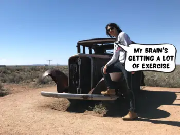 Meggie is wearing jeans, brown boots, and a white sweater. One foot is resting on the metal bumper of a rusty old car. Behind them is the desert and scrubby light green shrub plants in the desert. Big blue sky. Meggie says, "My brain's getting a lot of exercise." Petrified Forest National Park nearby Holbrook, Arizona, United States of America.