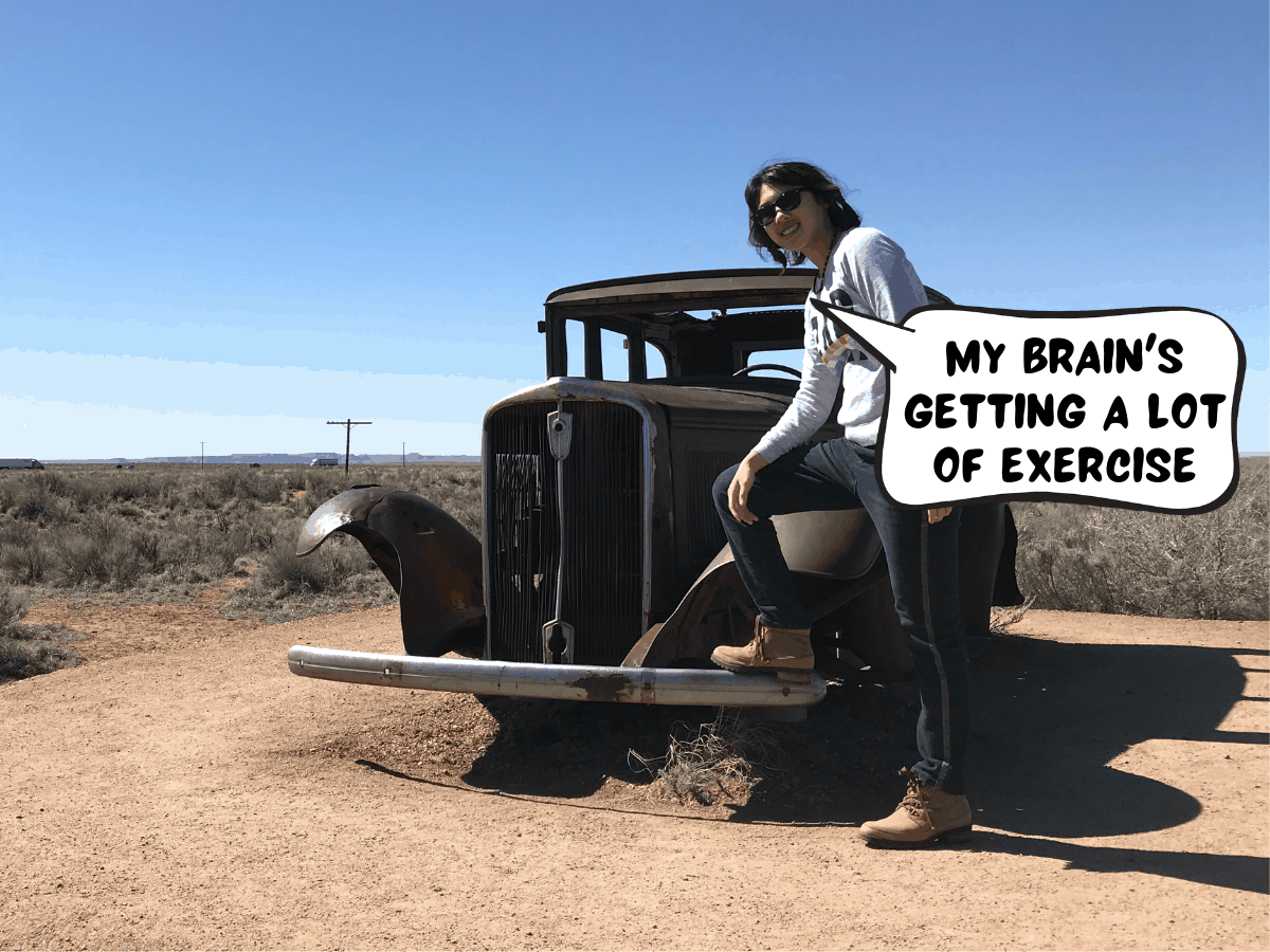 Meggie is wearing jeans, brown boots, and a white sweater. One foot is resting on the metal bumper of a rusty old car. Behind them is the desert and scrubby light green shrub plants in the desert. Big blue sky. Meggie says in a comic text bubble, "My brain's getting a lot of exercise." Petrified Forest National Park nearby Holbrook, Arizona, United States of America.