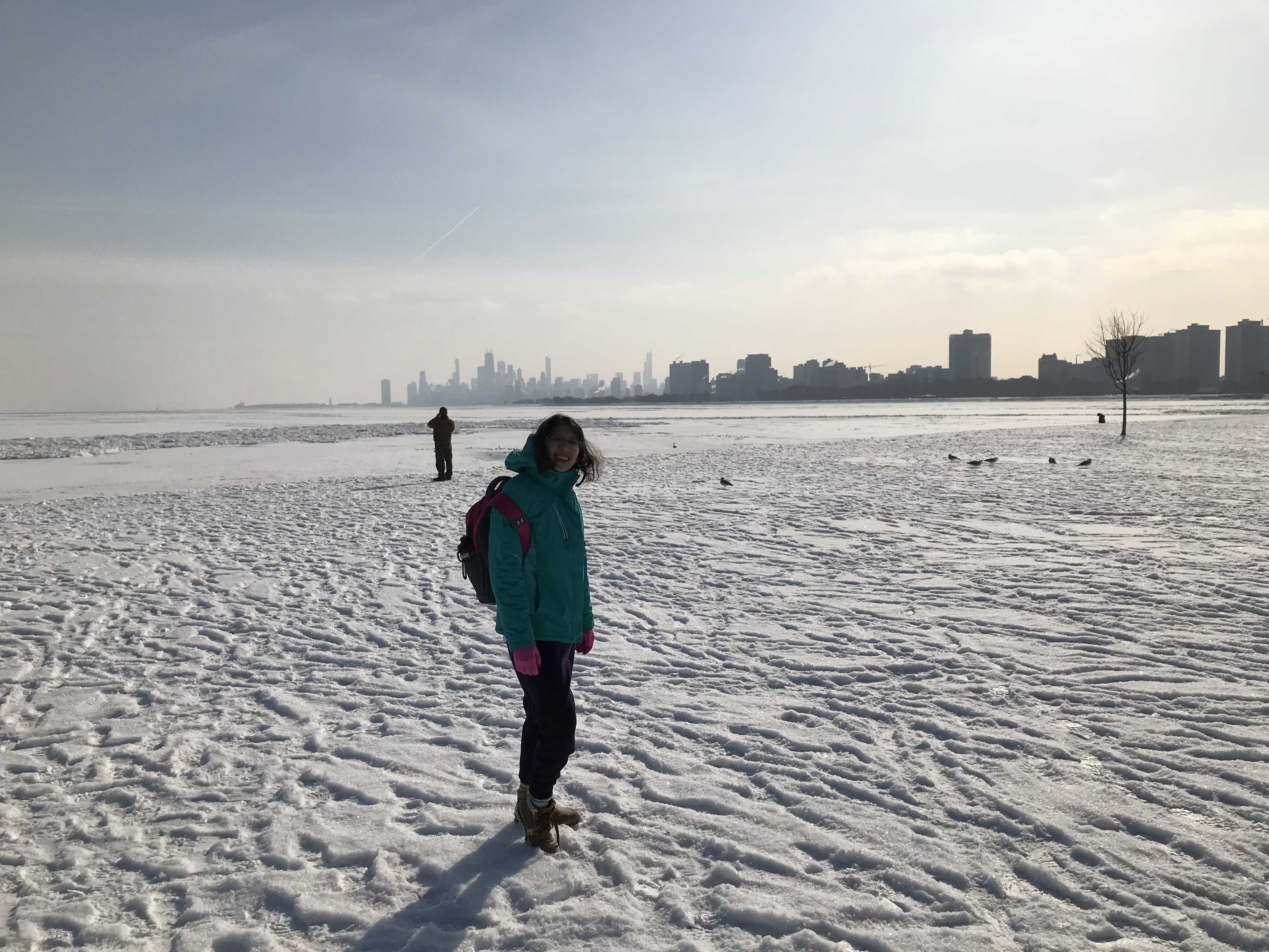 Lake Michigan and its coat of snow and ice, with the skyscraper backdrop of Chicago, Illinois, United States in the background. Meggie is wearing a thick blue jacket and brown boots. The sky is hazy white with a tint of blue.