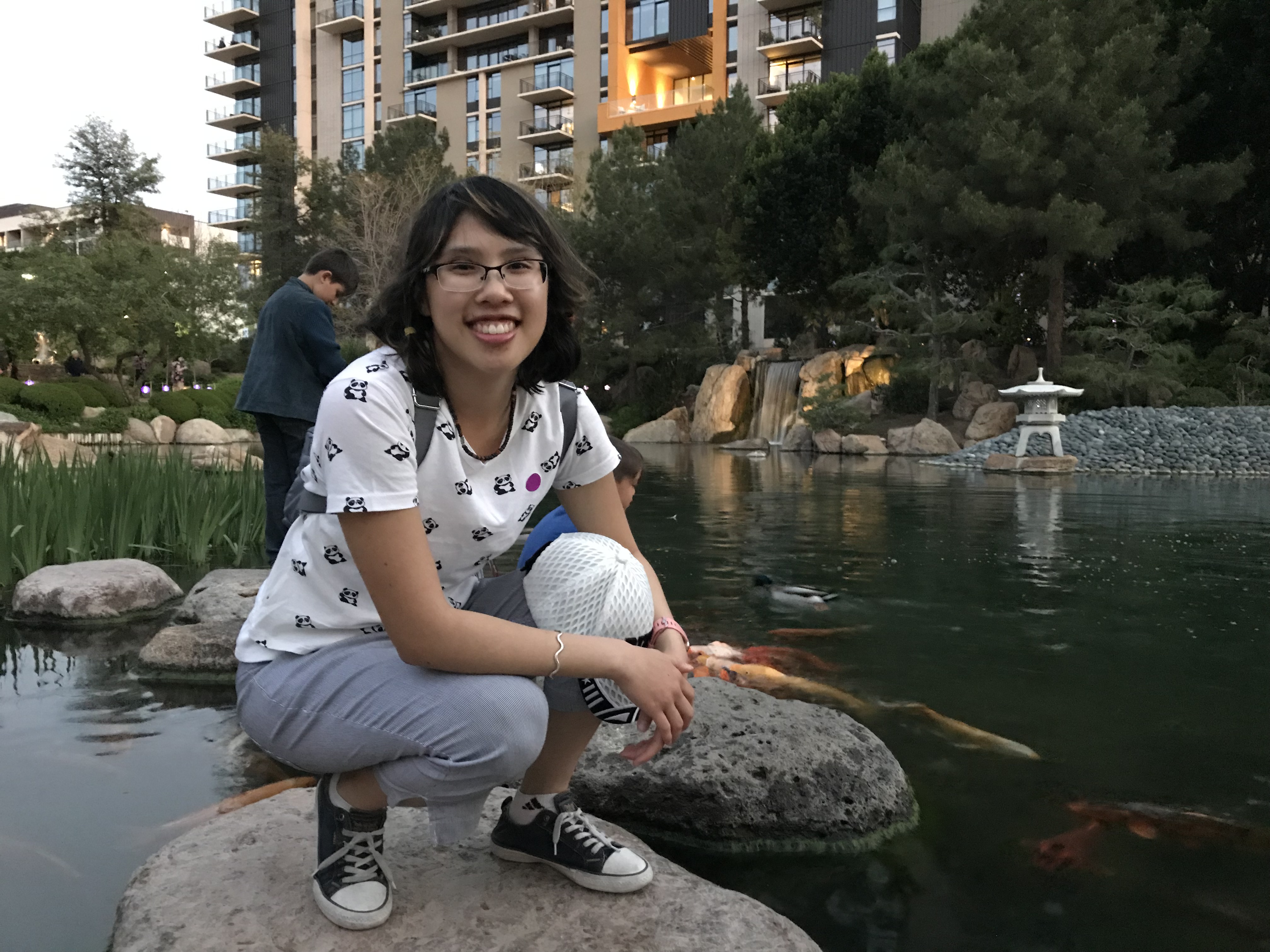 Meggie, with her white shirt and pants, is squatting on a rock in the lake of the Japanese Friendship Garden in Phoenix, Arizona, United States. Koi fish swim in the pond. Dark green trees and apartment buildings in the background.