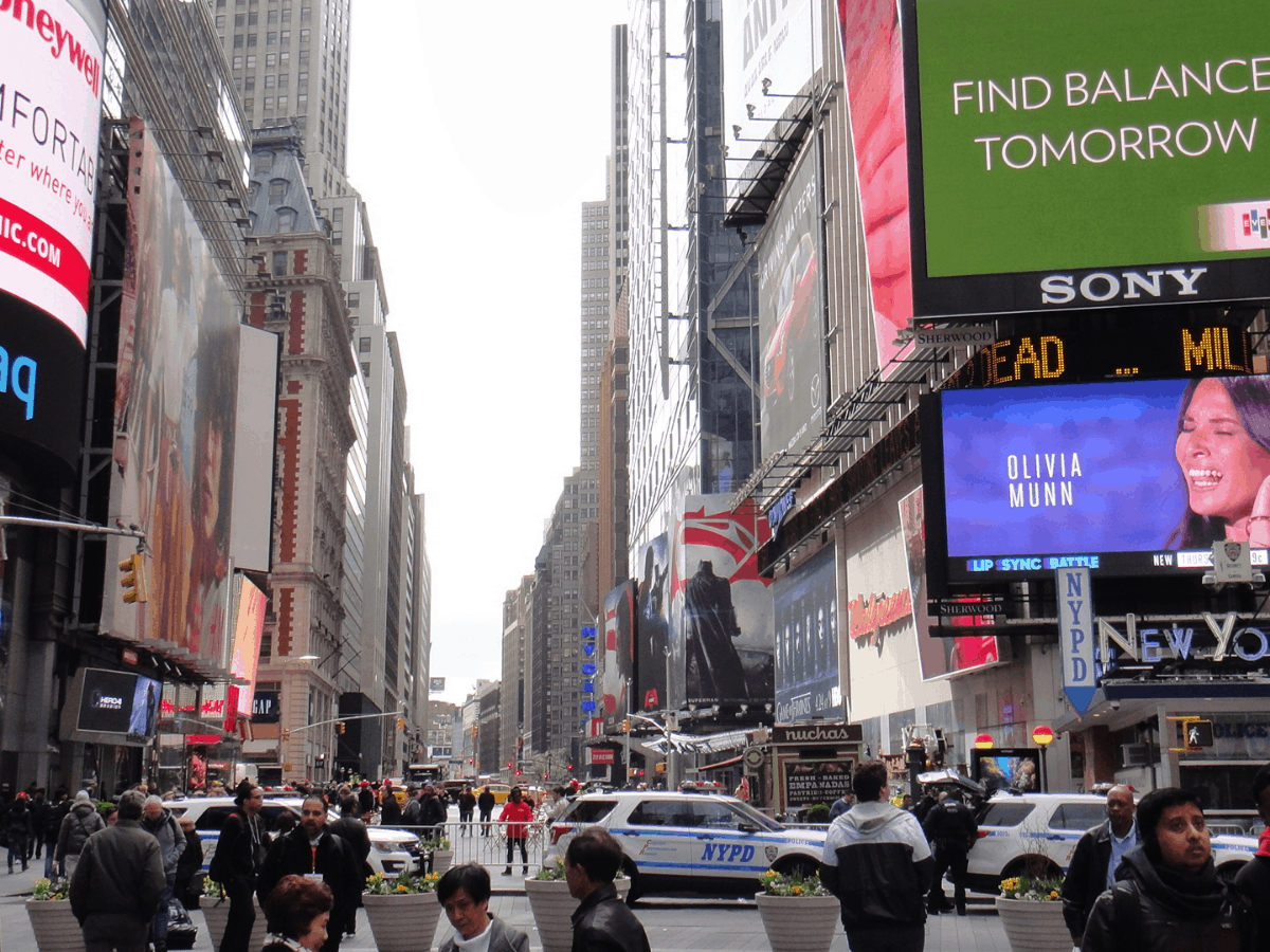 Times Square, Manhattan, New York City, New York State, United States of America. Lots of flashy lit-up billboards lining the street. Lots of tall buildings and skyscrapers. The cars are whizzing by. The scene is at day. The sky is cloudy and white. Crowds of people everywhere.