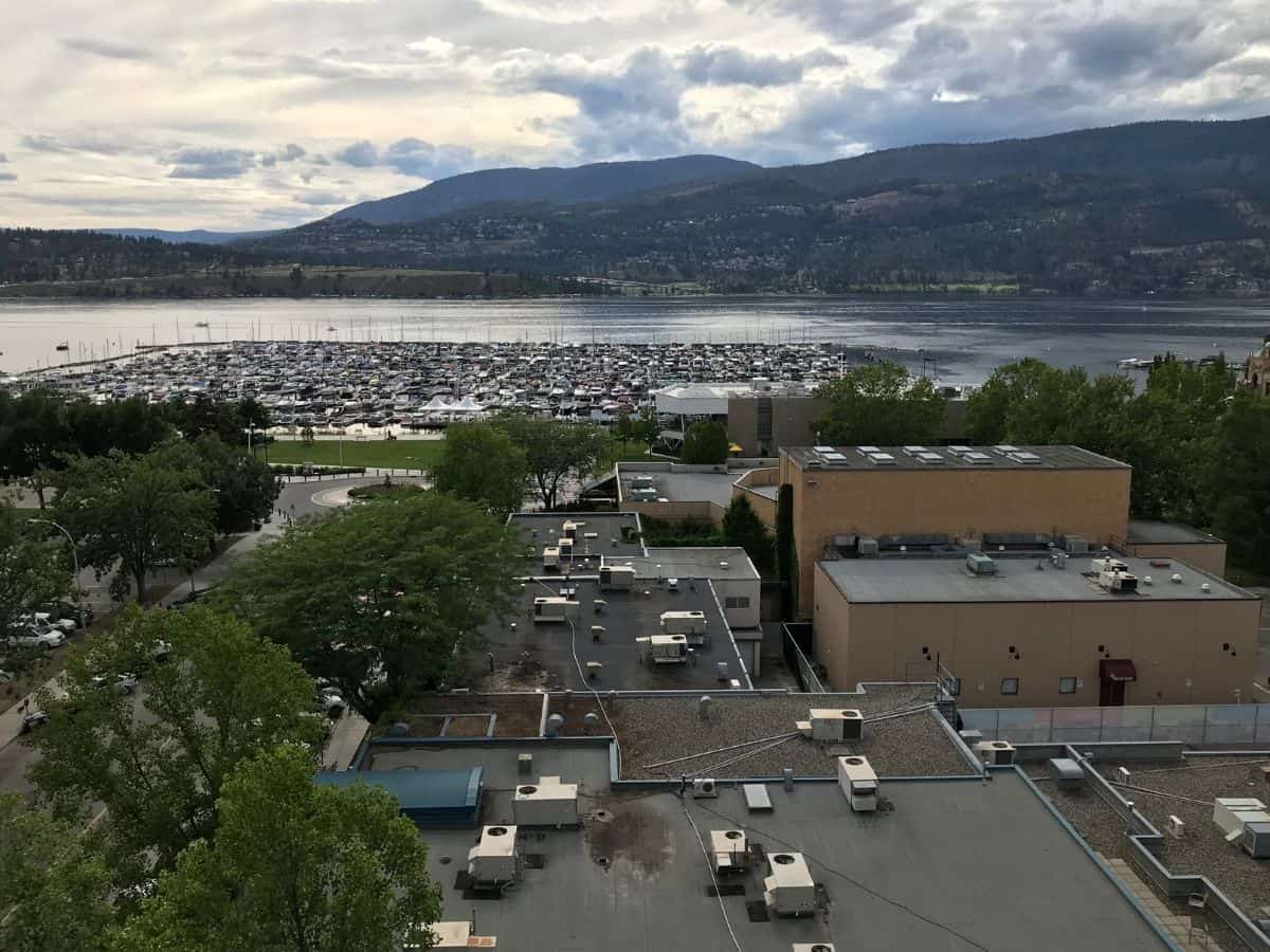 z wysokiego punktu widzenia brązowe budynki i zielone zadrzewione ulice kończą się w marinie pełnej zaparkowanych żaglówek na brzegu jeziora Okanagan. Niebo jest pochmurne i ponure, rzucając ciemny cień na krajobraz w Kelowna, Kolumbia Brytyjska, Kanada