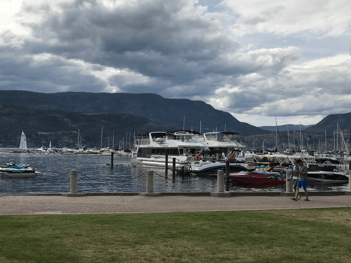 At the ground level, sailboats are docked at the downtown Marina. A couple is walking on the sidewalk along the dock. The sky is cloudy and gloomy. Kelowna, British Columbia, Canada