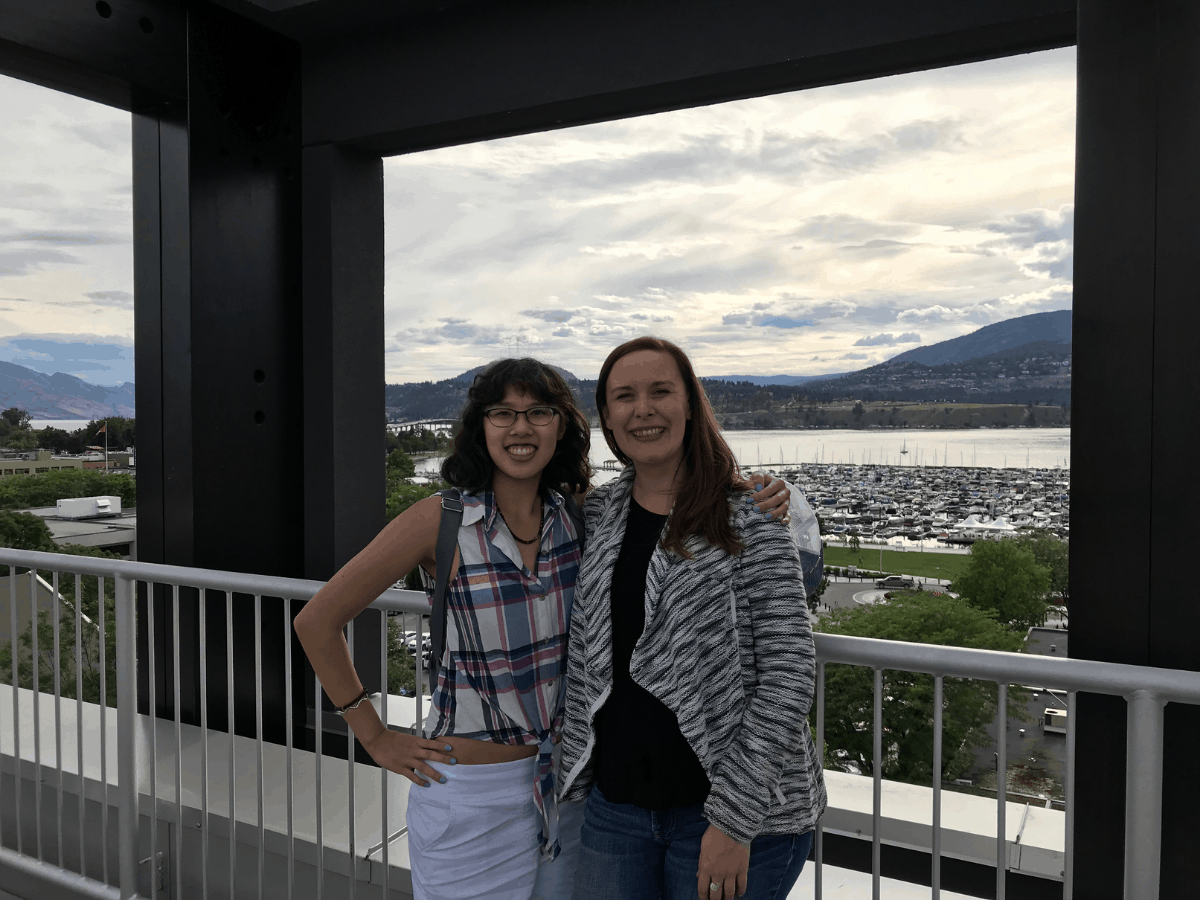 Meggie and Emma/Bambalou stand at Lane Merrifield/Billybob's Perch Cafe (on top of the Okanagan Centre for Innovation) overlooking the Kelowna downtown marina, Okanagan Lake, and surrounding mountains. Sky is cloudy. Kelowna, British Columbia, Canada