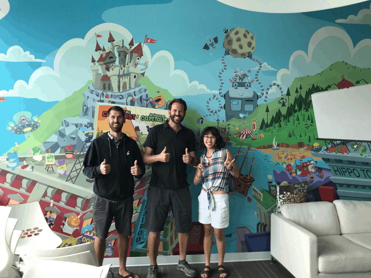 Meggie is with Lance Priebe/RocketSnail and Tristan Rattink at the Hyper Hippo Productions/Games headquarters in Kelowna, British Columbia, Canada. They are all doing a thumbs-up, and standing in front of a colorful, playful painted mural full of hippo characters.