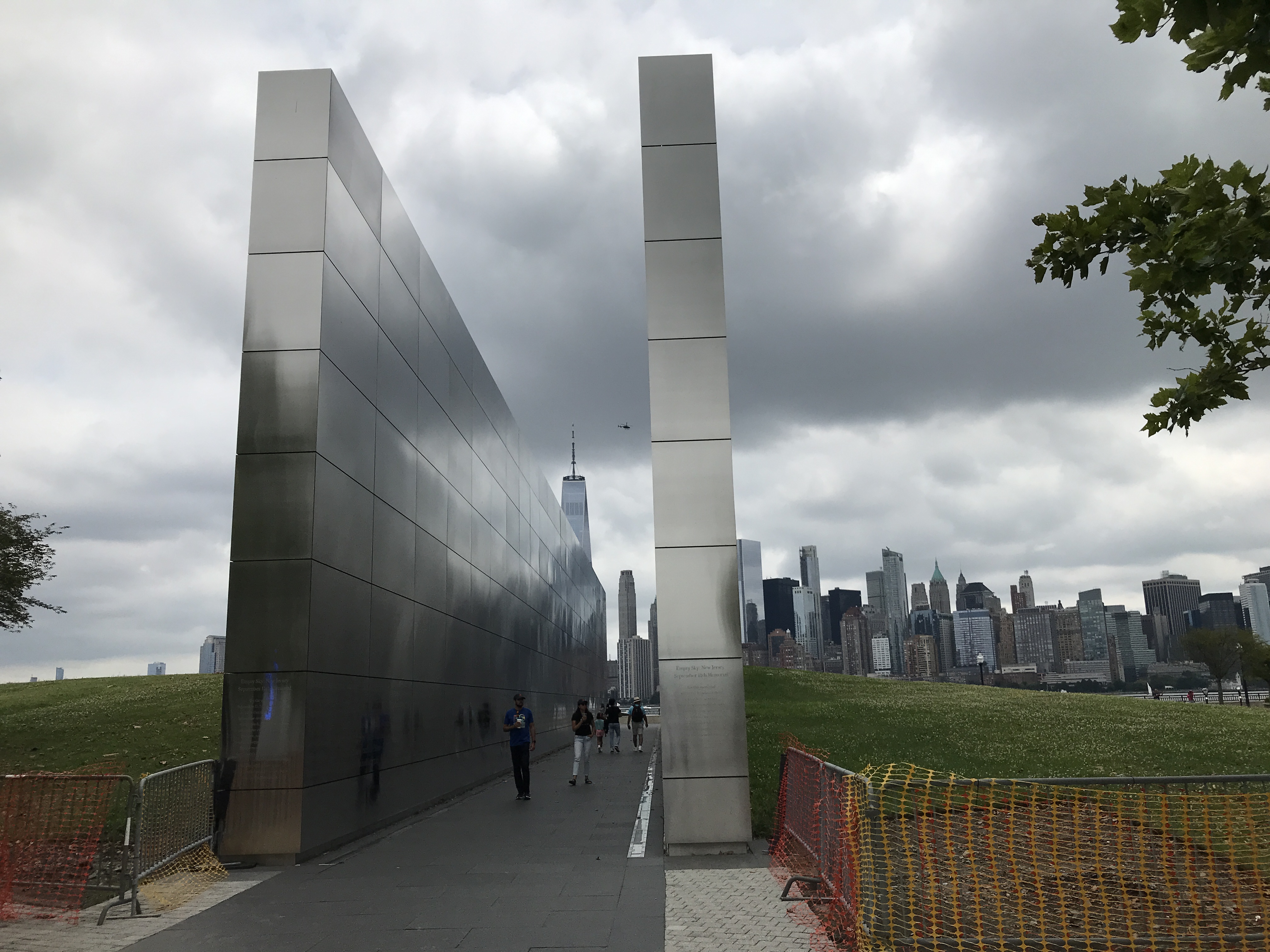 The Empty Sky Memorial has two long rectangular stainless steel walls with the New York City skyscrapers in the background. Sky has dark cloud coverage. Jersey City, New Jersey, United States