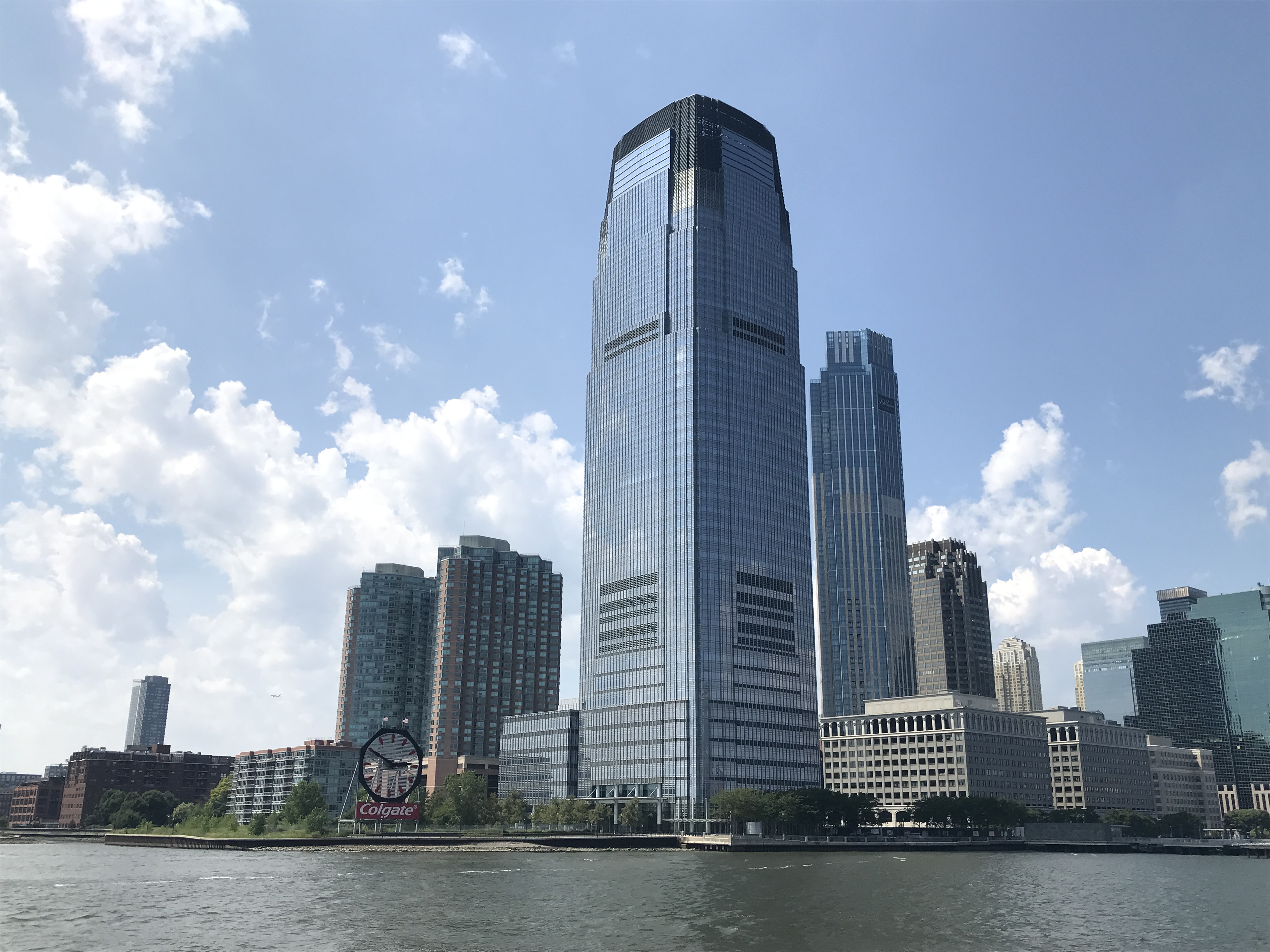 The extremely tall and glossy Goldman Sachs Building and the Colgate Clock sitting at the base of the building can be seen from the Hudson River. Sky is party sunny with some clouds. Jersey City, New Jersey, United States