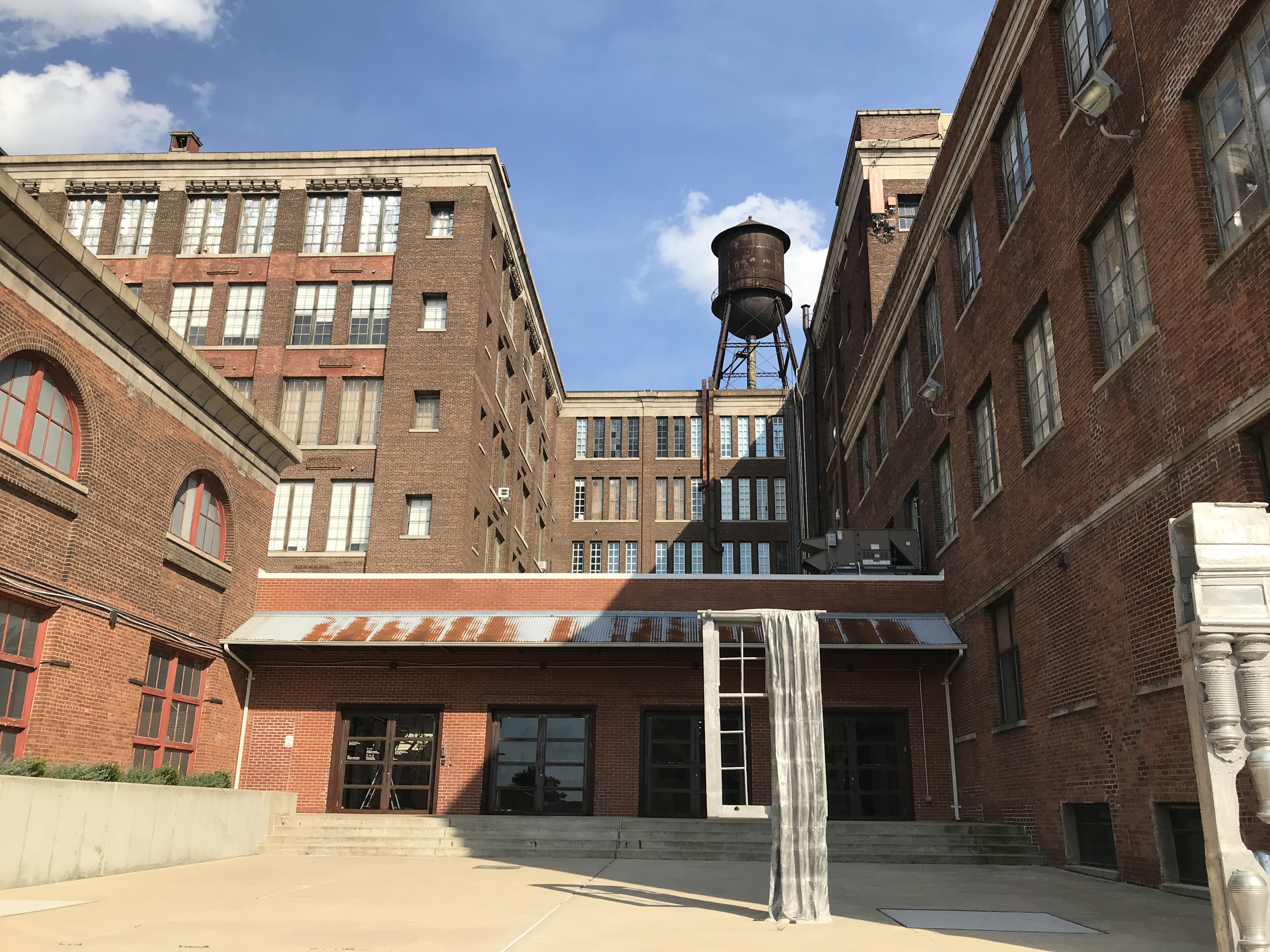 MANA Contemporary's dark brown and red bricked buildings give off an industrial, old feeling. There is even a black water tower in the background. Jersey City, New Jersey, United States. Sky is mostly clear blue with some clouds.