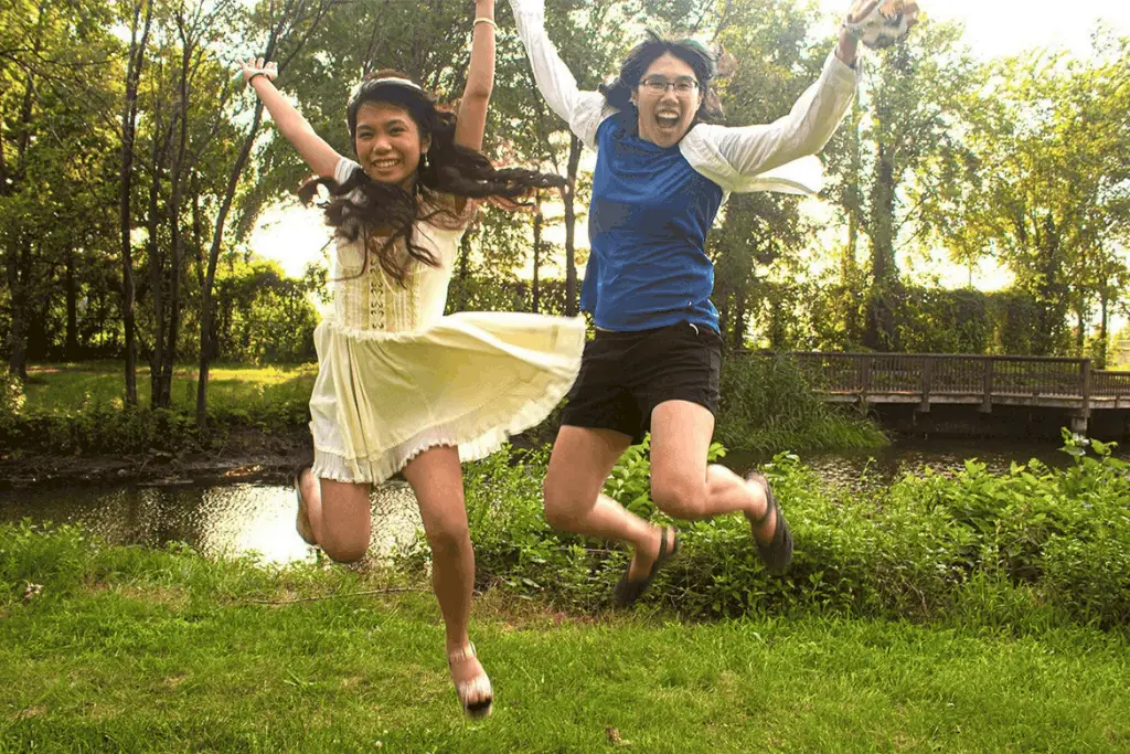 Meggie, dressed in a blue shirt and white overshirt, and Athena, dressed in a cute white dress, are grinning and jumping in the air in a nature park with green trees, a creek, and grass. Lincoln Park, Jersey City, New Jersey, United States of America