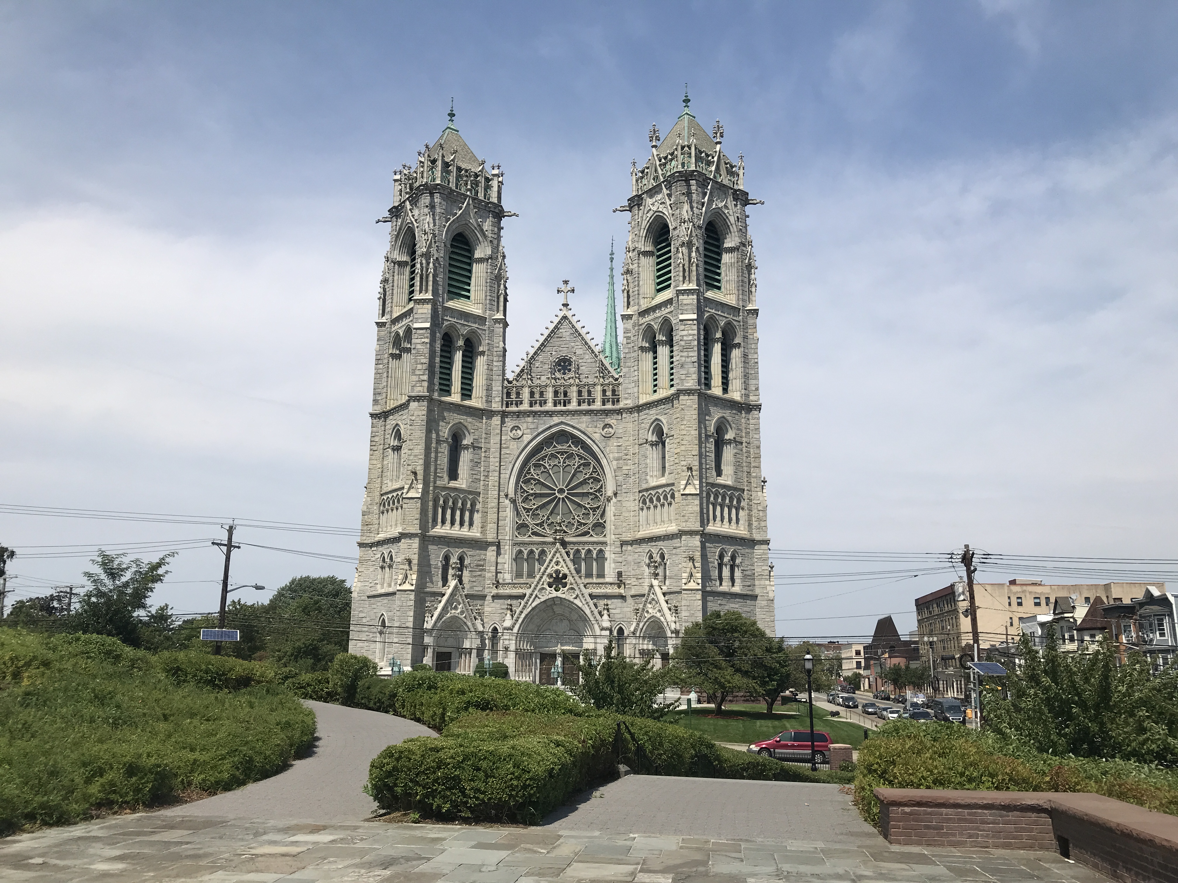 The grey, tall towers of the Cathedral Basilica of the Sacred Heart. Many long windows adorn the church. The ground is concrete with some green bushes. Newark, New Jersey, United States of America.