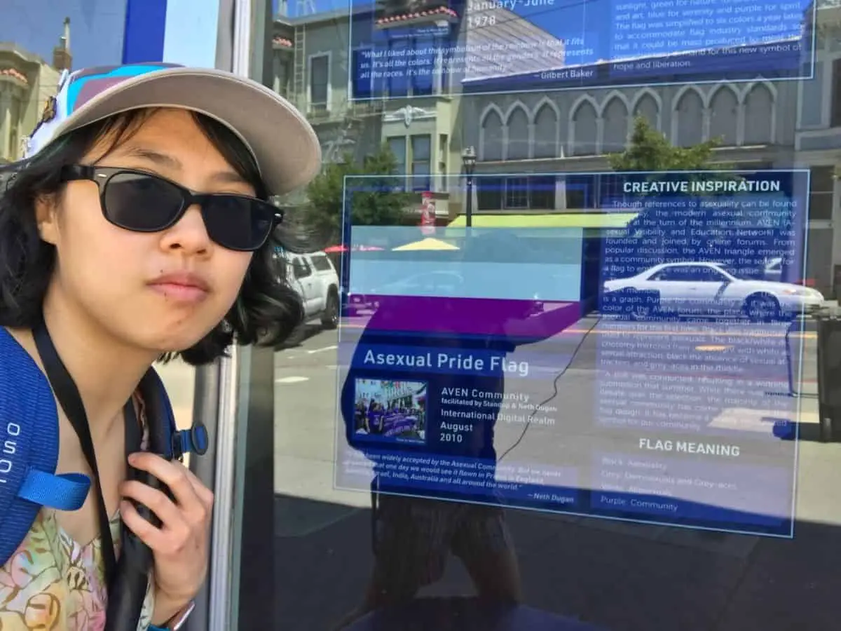 Meggie is standing solemnly in front of the window display of the black, gray, white, and purple asexual flag. The Castro District, San Francisco, California, United States of America