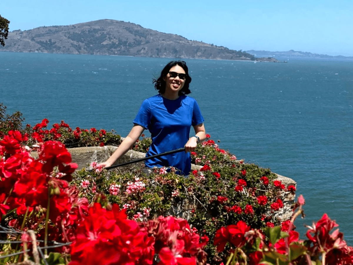 Meggie is grinning amongst red rose bushes overlooking the dark blue waters of the San Francisco Bay on Alcatraz Island, San Francisco, California, United States of America.